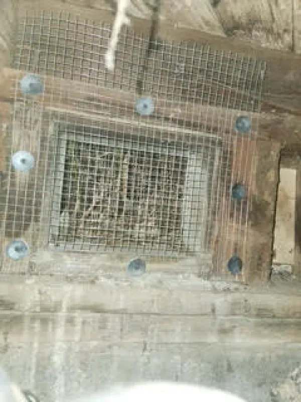 A wire mesh screen is secured over a rectangular wall vent using several large metal washers and screws.