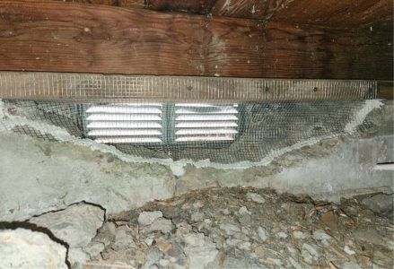 A rectangular crawl space vent with wire mesh installed in a crumbling concrete foundation wall below a wooden joist.