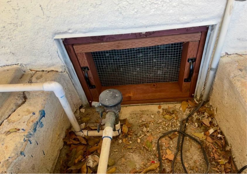 A wooden-framed window well cover with wire mesh installed over an exterior foundation vent near pipes and ground debris.