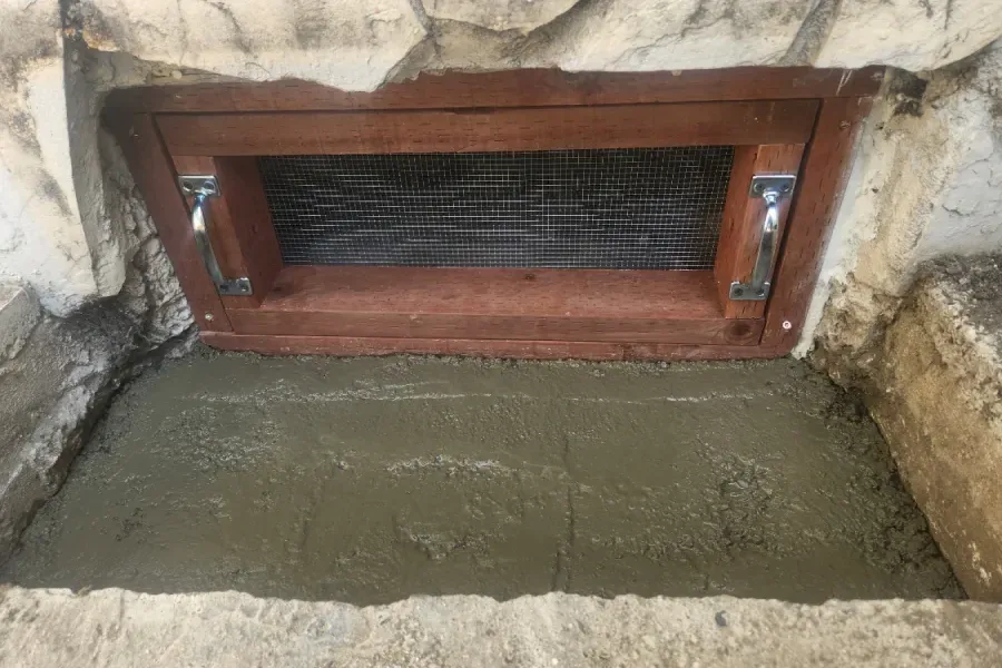 A rectangular wooden framed vent with a wire mesh screen, set into a stone wall above a freshly poured concrete patch.