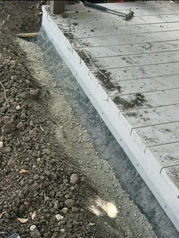 A trench filled with gravel runs along the side of a grey wooden deck, with exposed foundation and a hammer visible.