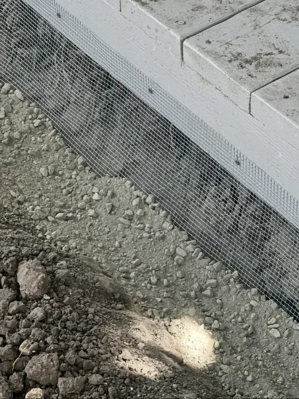 A close-up view of a metal mesh screen installed along the base of a light grey structure, bordering loose gravel and soil.