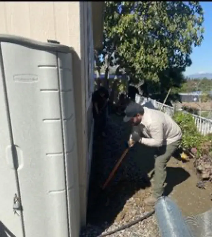 A person in a light shirt and dark cap uses a shovel to dig a trench along the side of a building next to a plastic shed.