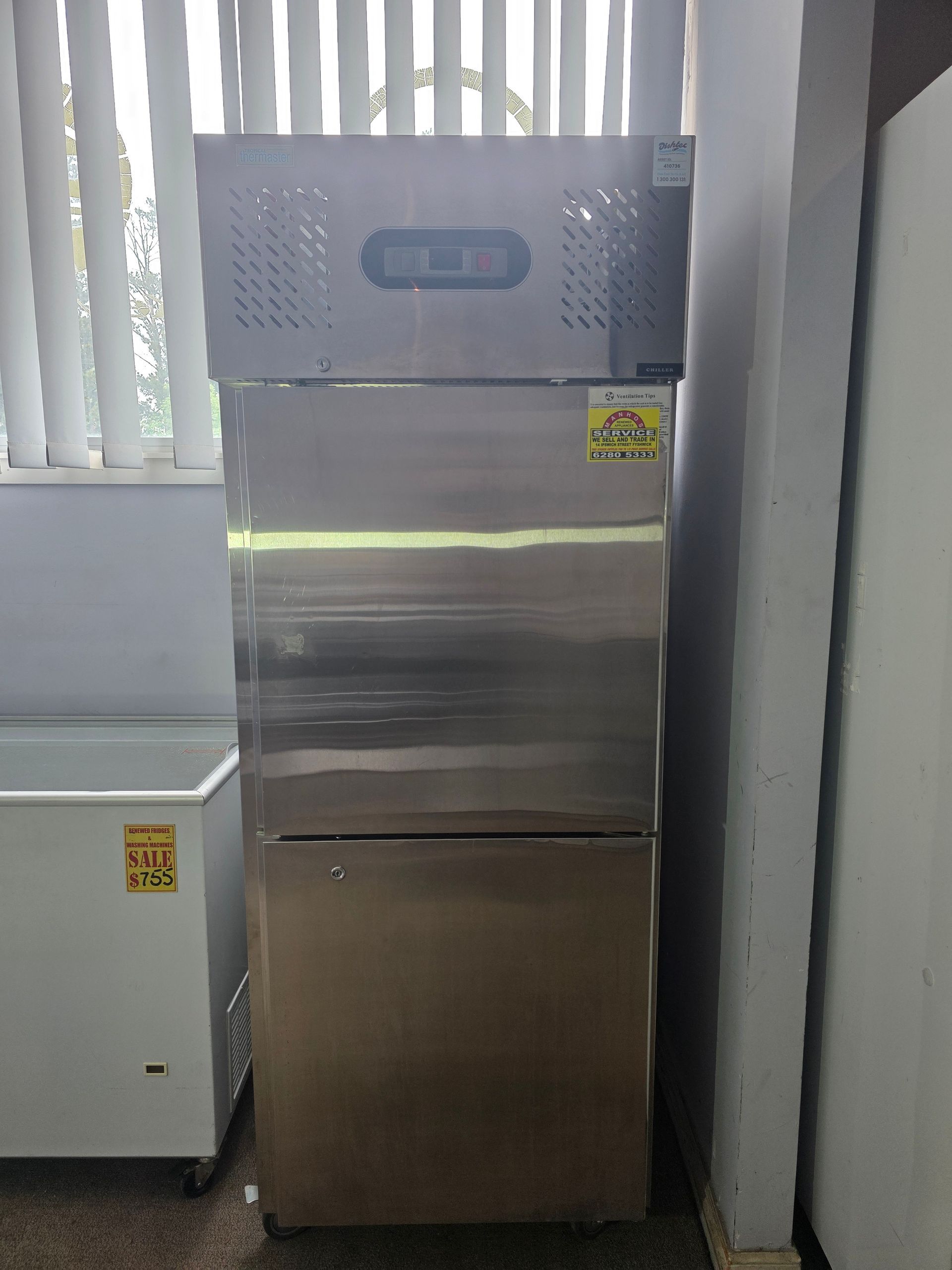Stainless steel commercial refrigerator, two-door, indoors near a window and white chest freezer.
