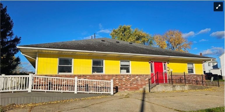Yellow building with red door, brick facade, white fence, and a blue sky.