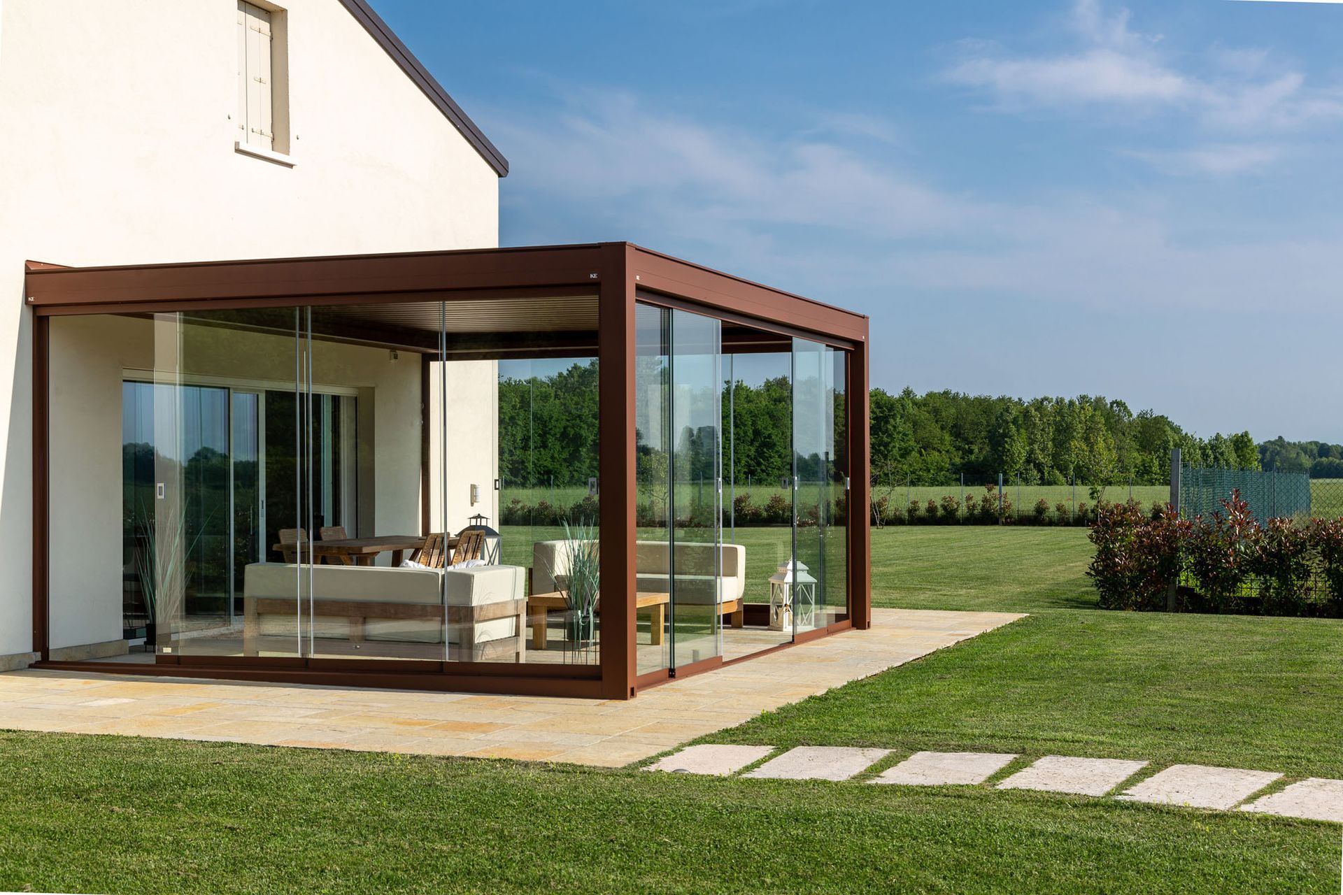 Veranda con pareti in vetro annessa a una casa bianca con struttura marrone, arredata e dotata di prato erboso.