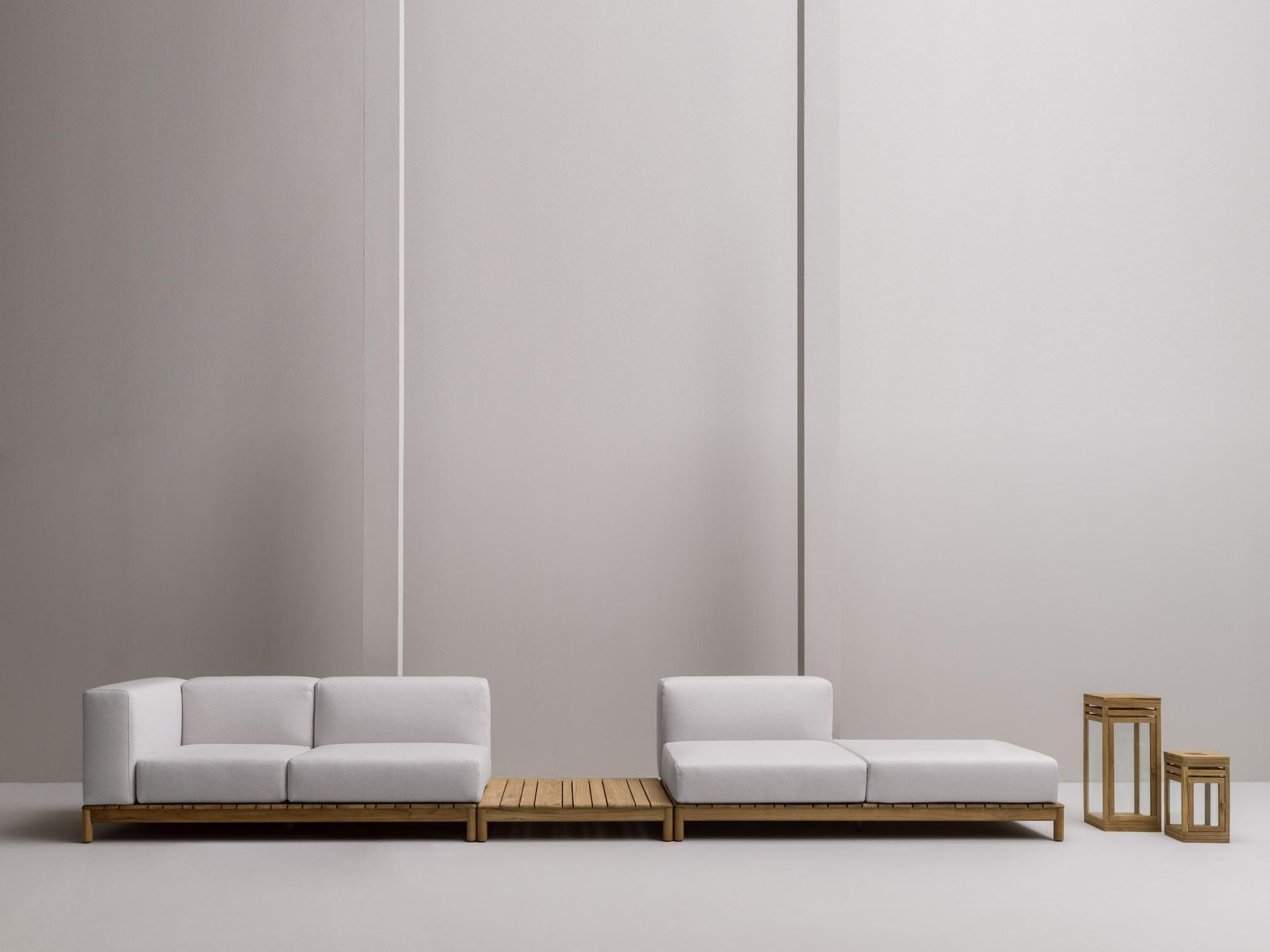 Divano componibile bianco con base in legno su sfondo grigio chiaro, accanto a due lanterne in legno.