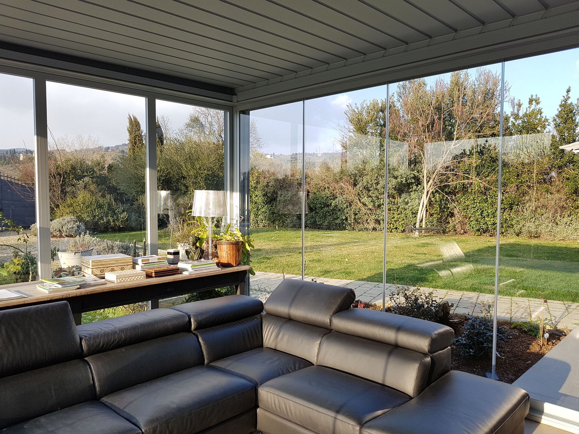 Veranda con pareti in vetro e divano in pelle nera, con vista su un cortile verde e alberi.