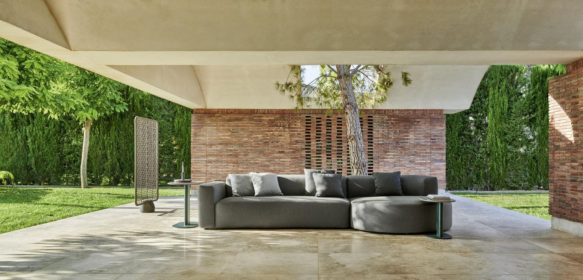 Area lounge esterna coperta con divano grigio, muro di mattoni e vegetazione lussureggiante.