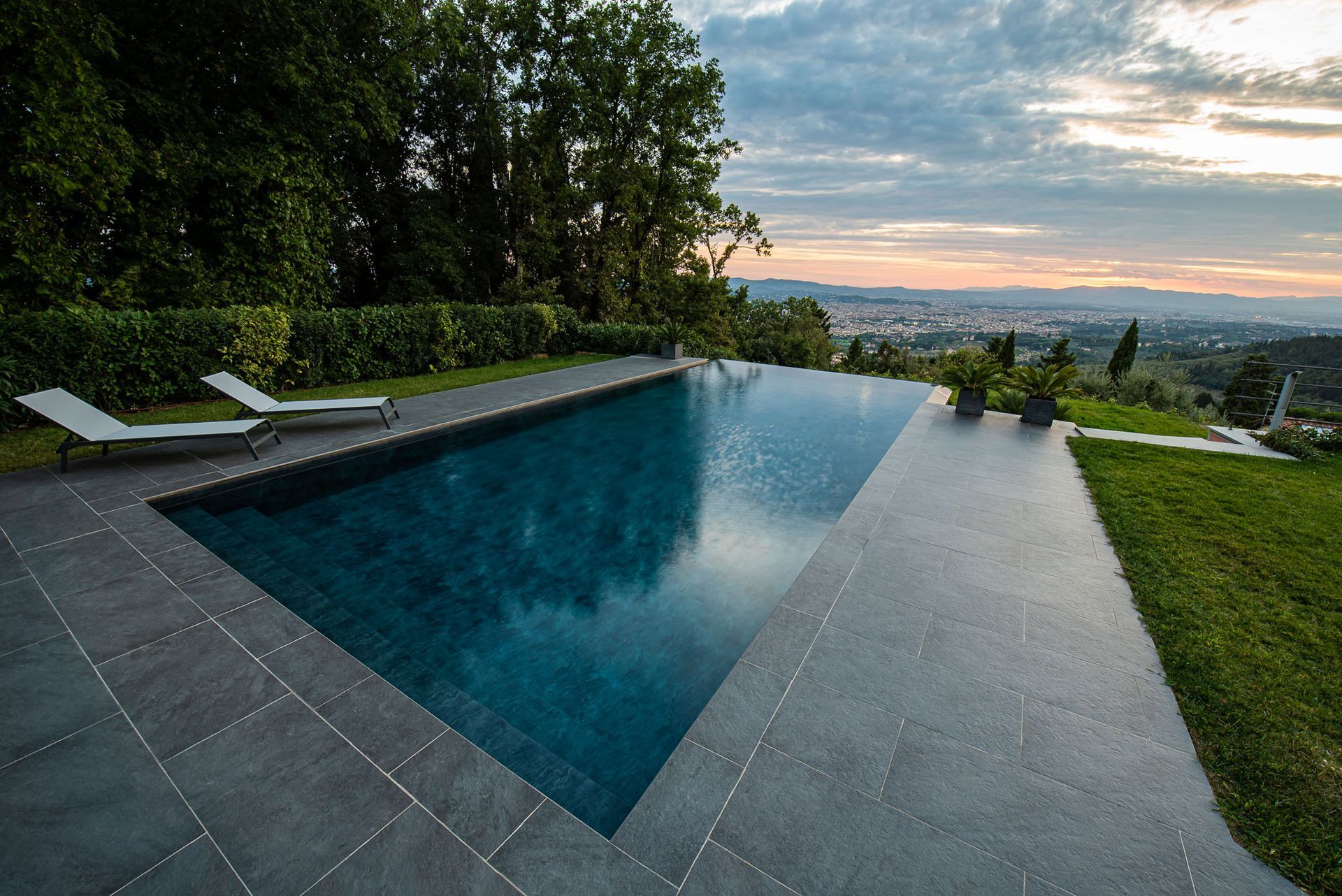 Piscina a sfioro con vista sulla valle al tramonto, dotata di sedie a sdraio e vegetazione verde.