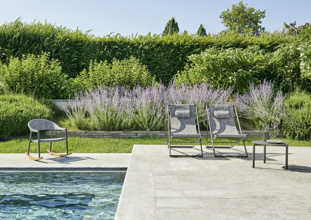 Scena a bordo piscina con mobili da esterno grigi: sedie e un tavolino. Verde lussureggiante sullo sfondo.