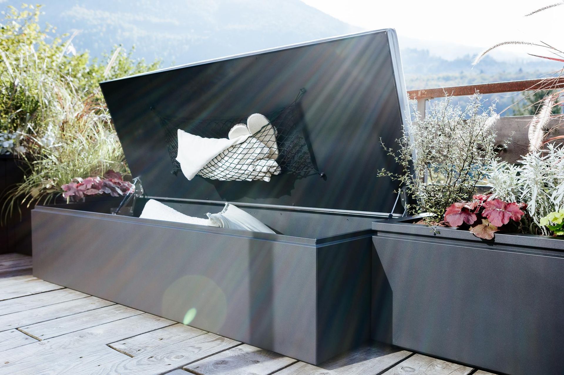 Contenitore da esterno con coperchio aperto, cuscini visibili e sfondo montano.