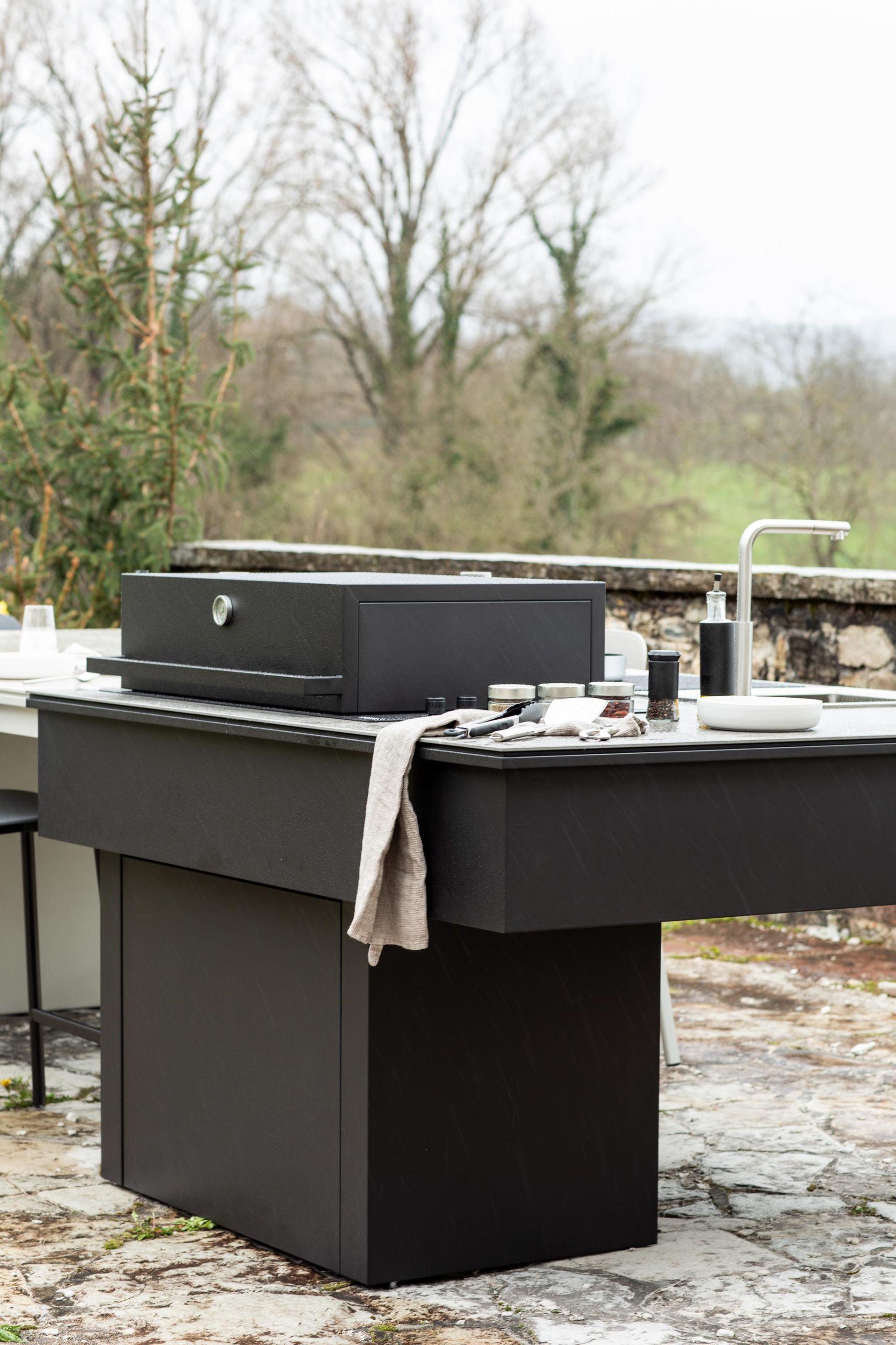 Isola da cucina esterna nera con grill e lavello, incastonata tra gli alberi.