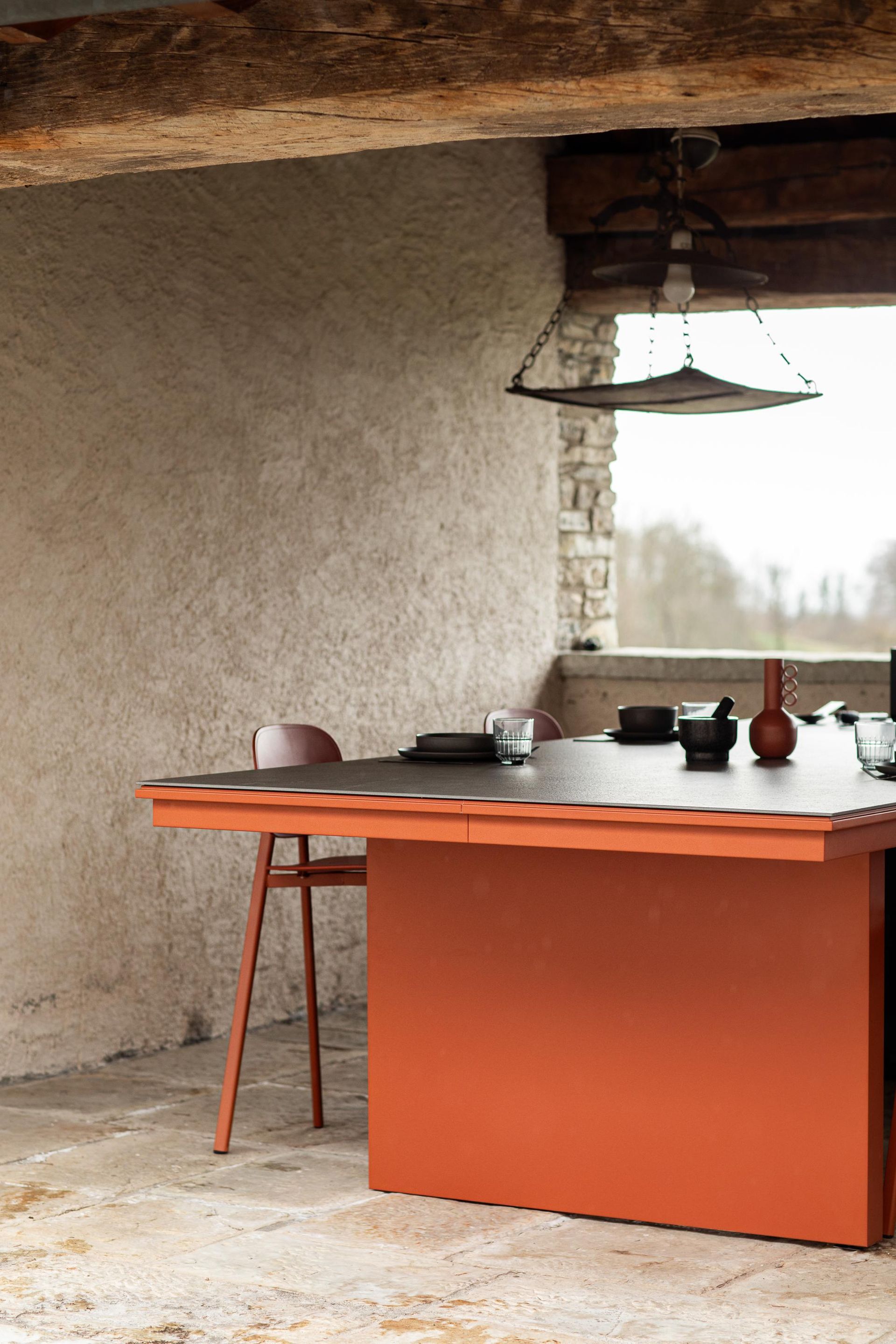 Tavolo e sedie in terracotta su un patio esterno coperto; muro in pietra, travi in ​​legno, luce dall'alto, stoviglie.