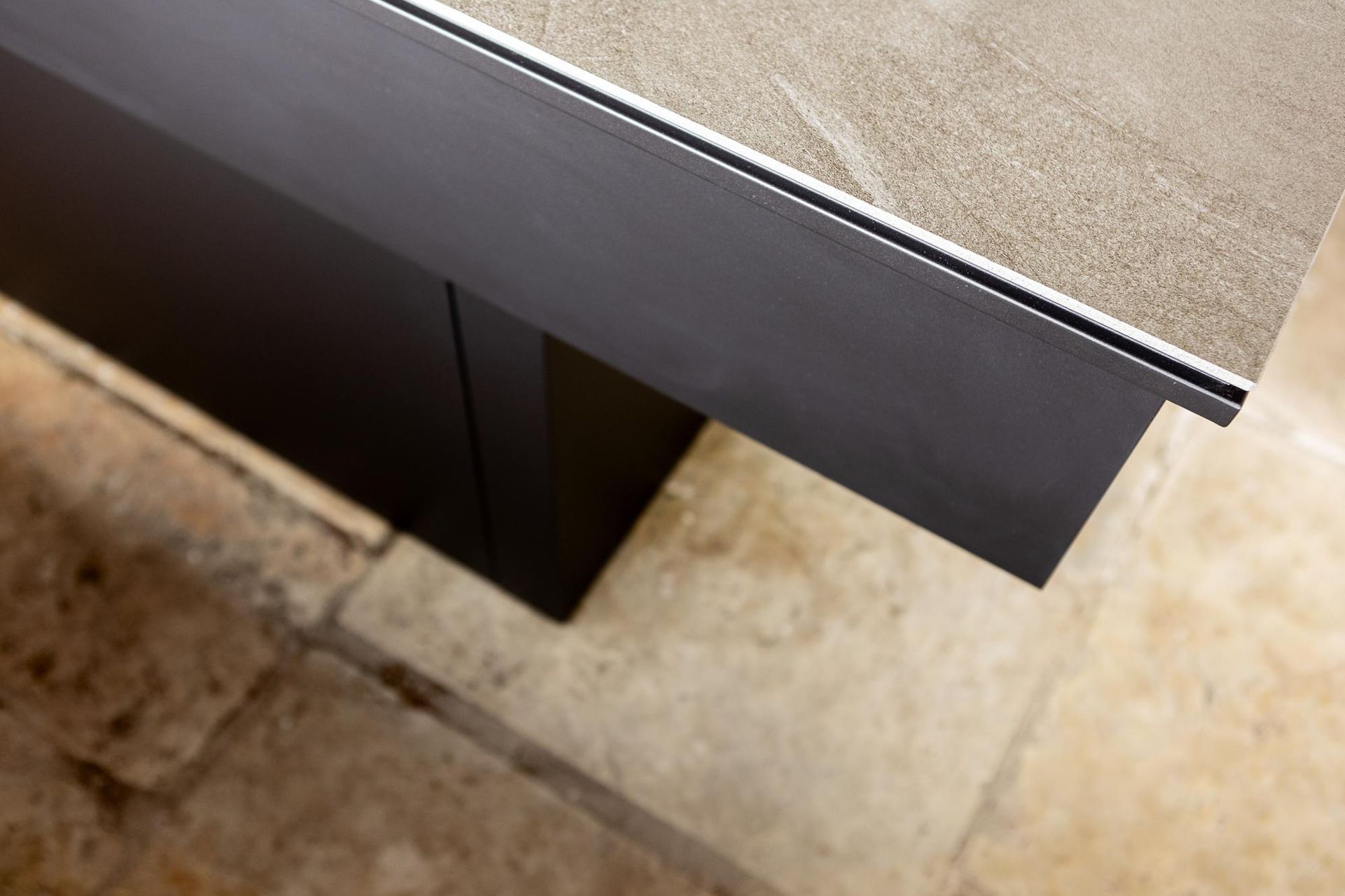 Tavolo grigio scuro con piano in pietra e finiture in metallo, appoggiato su un pavimento in pietra.