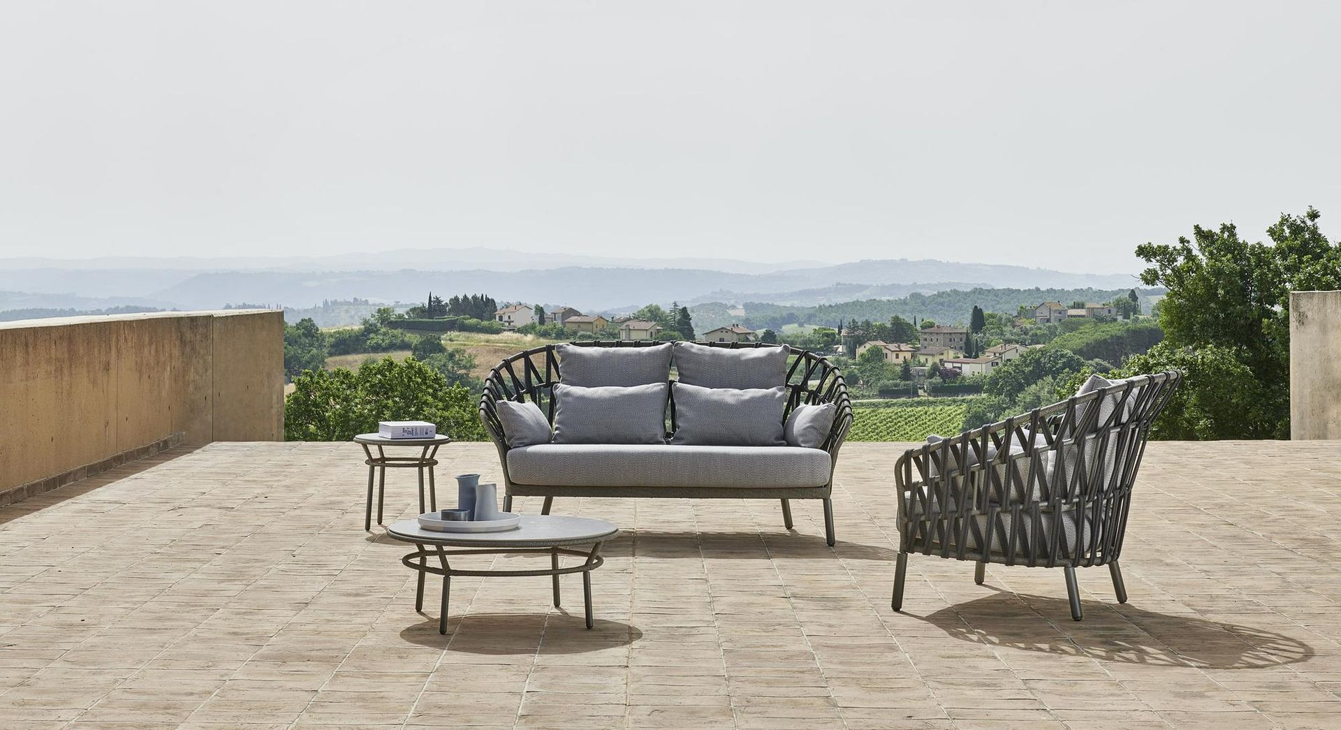 Set da esterno per patio con divano, sedia e tavoli con vista sul paesaggio. Cuscini grigi e strutture intrecciate.
