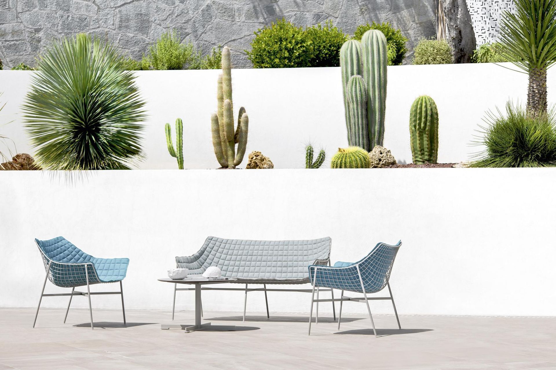 Set di mobili da giardino con sedie intrecciate blu e un divano di fronte a cactus e un muro bianco.