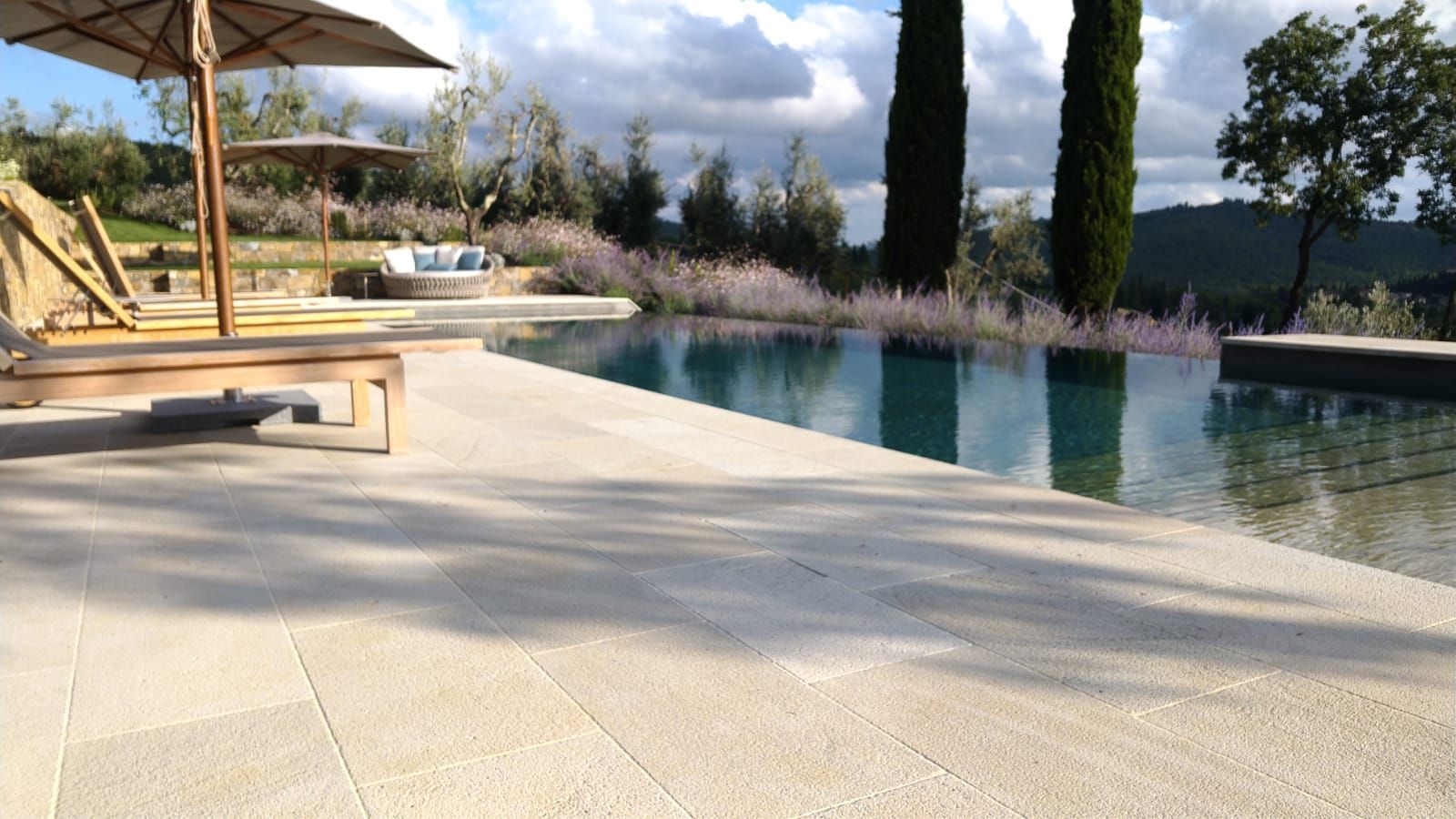 Scena a bordo piscina con patio in pietra, acqua, ombrelloni e paesaggio toscano sotto un cielo nuvoloso.