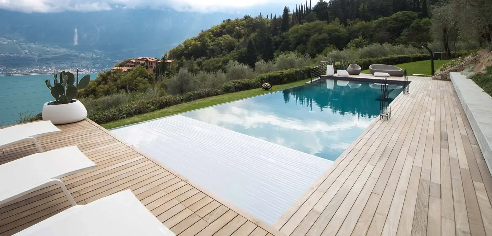 Piscina con acqua, circondata da una terrazza in legno e verde, con vista sul lago e sulle montagne.