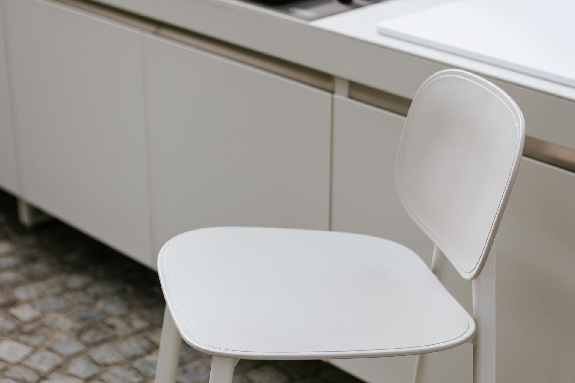 Sedia bianca davanti ai mobili bianchi della cucina, su un pavimento piastrellato.