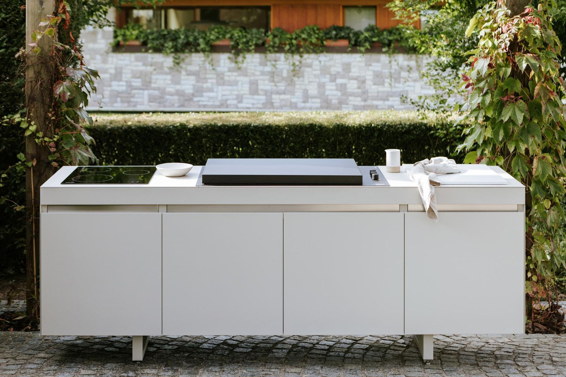 Isola da cucina bianca da esterno con piano cottura e mobili integrati, con vista sul verde e su un muro di mattoni.