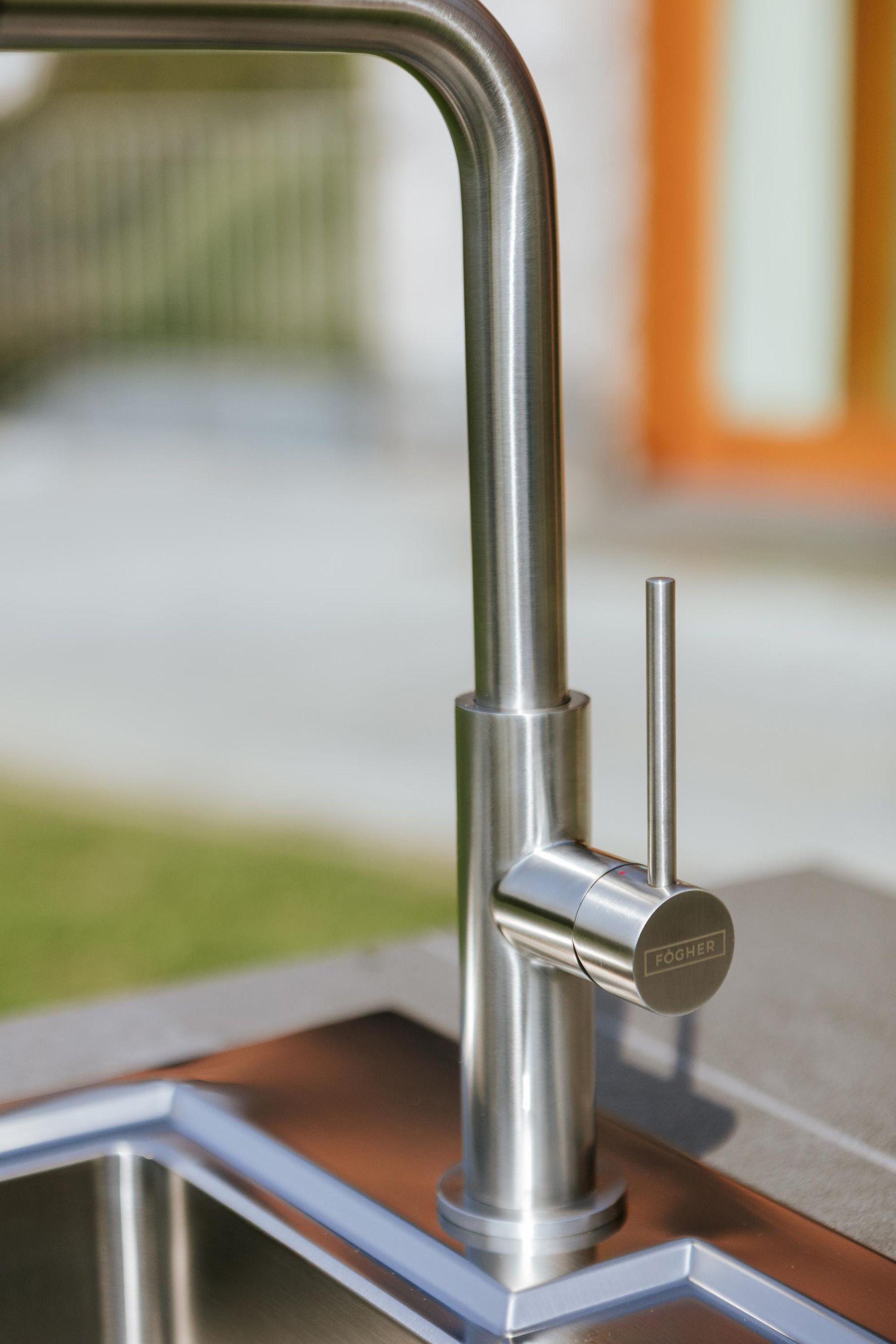 Rubinetto da cucina in acciaio inossidabile sopra un lavello; all'aperto con vegetazione e uno sfondo arancione.