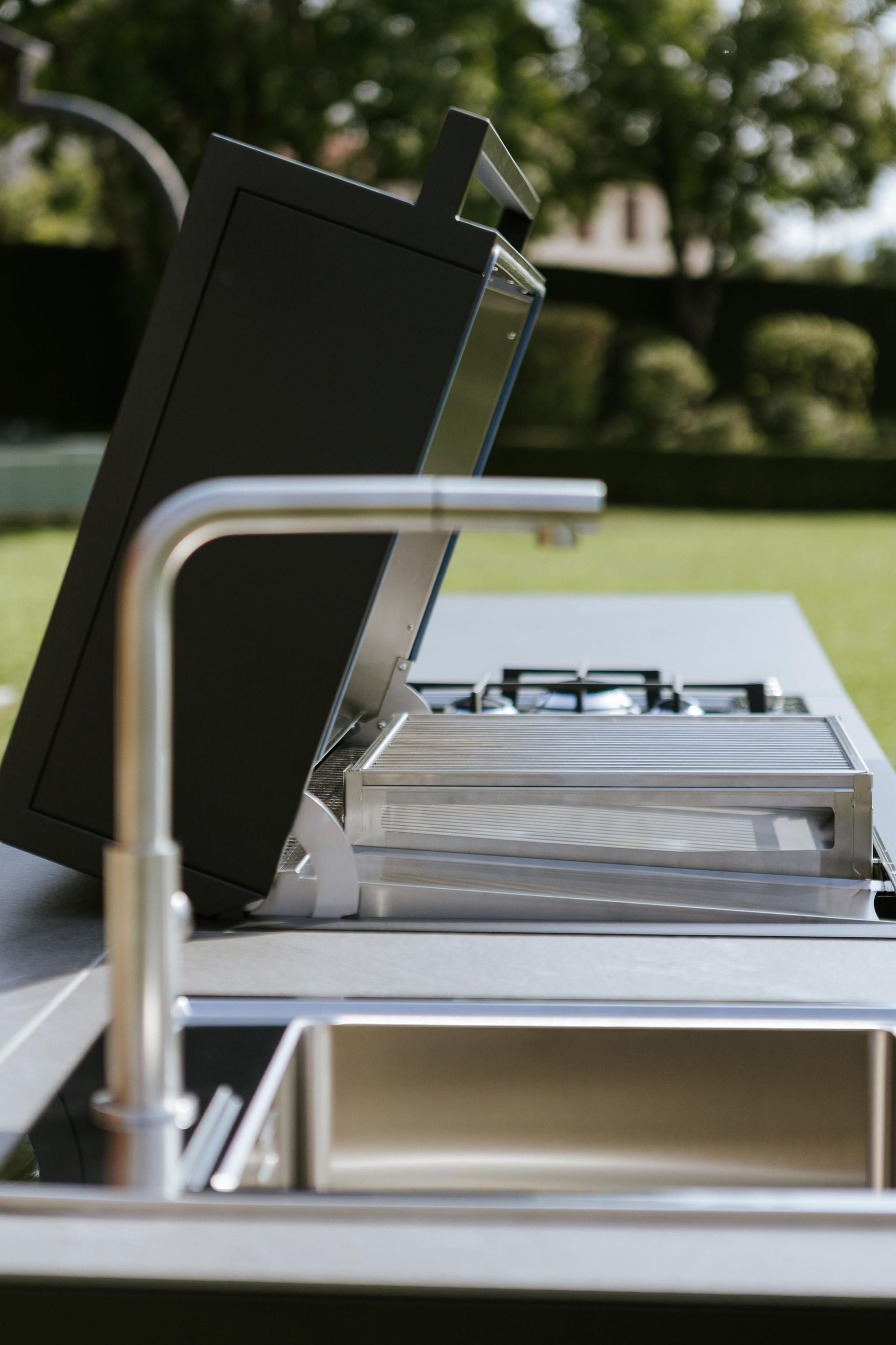 Cucina da esterno con coperchio rialzato sopra la griglia, lavello in acciaio inox e rubinetto.