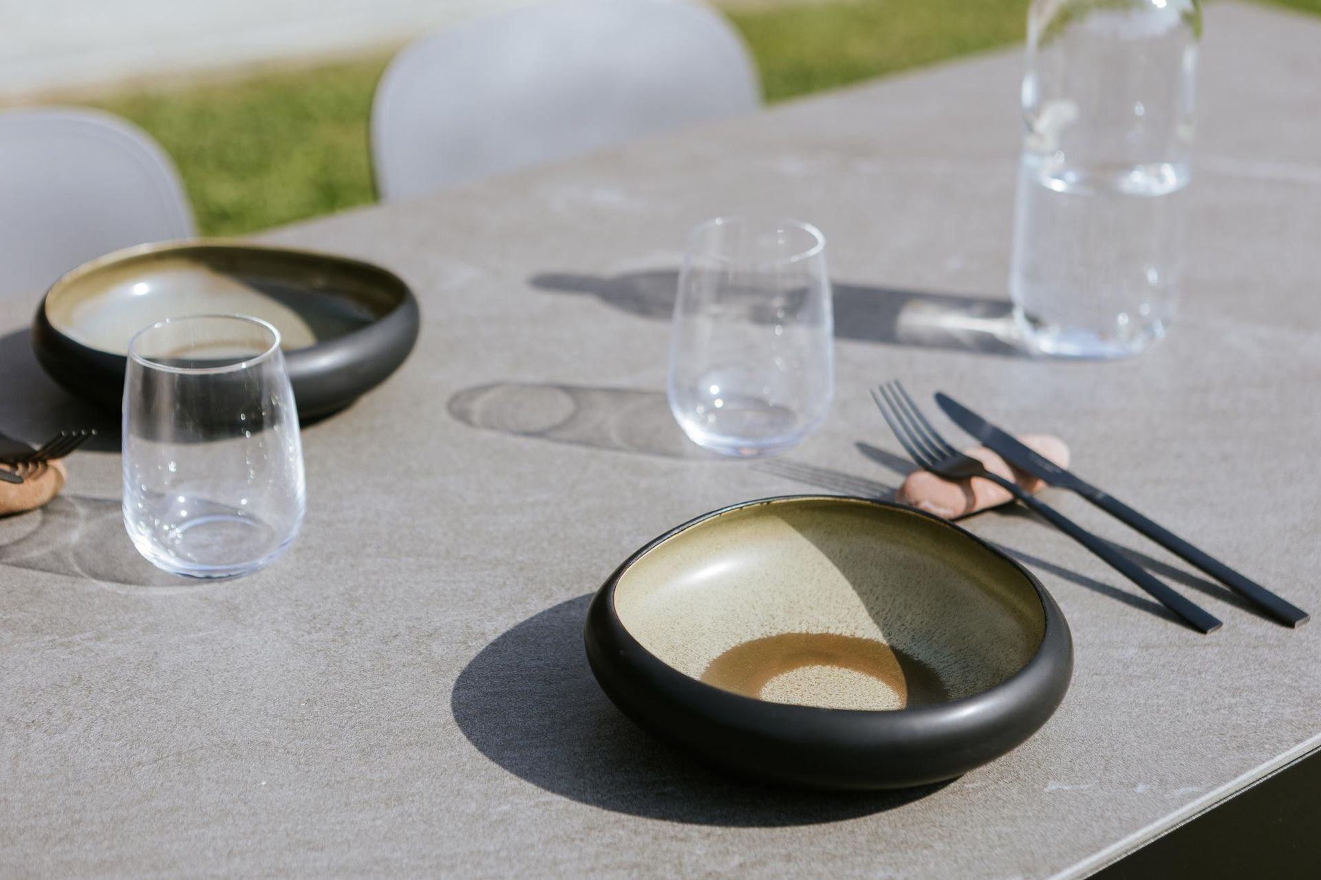 Tavolo apparecchiato per due all'aperto con ciotole, bicchieri e posate.