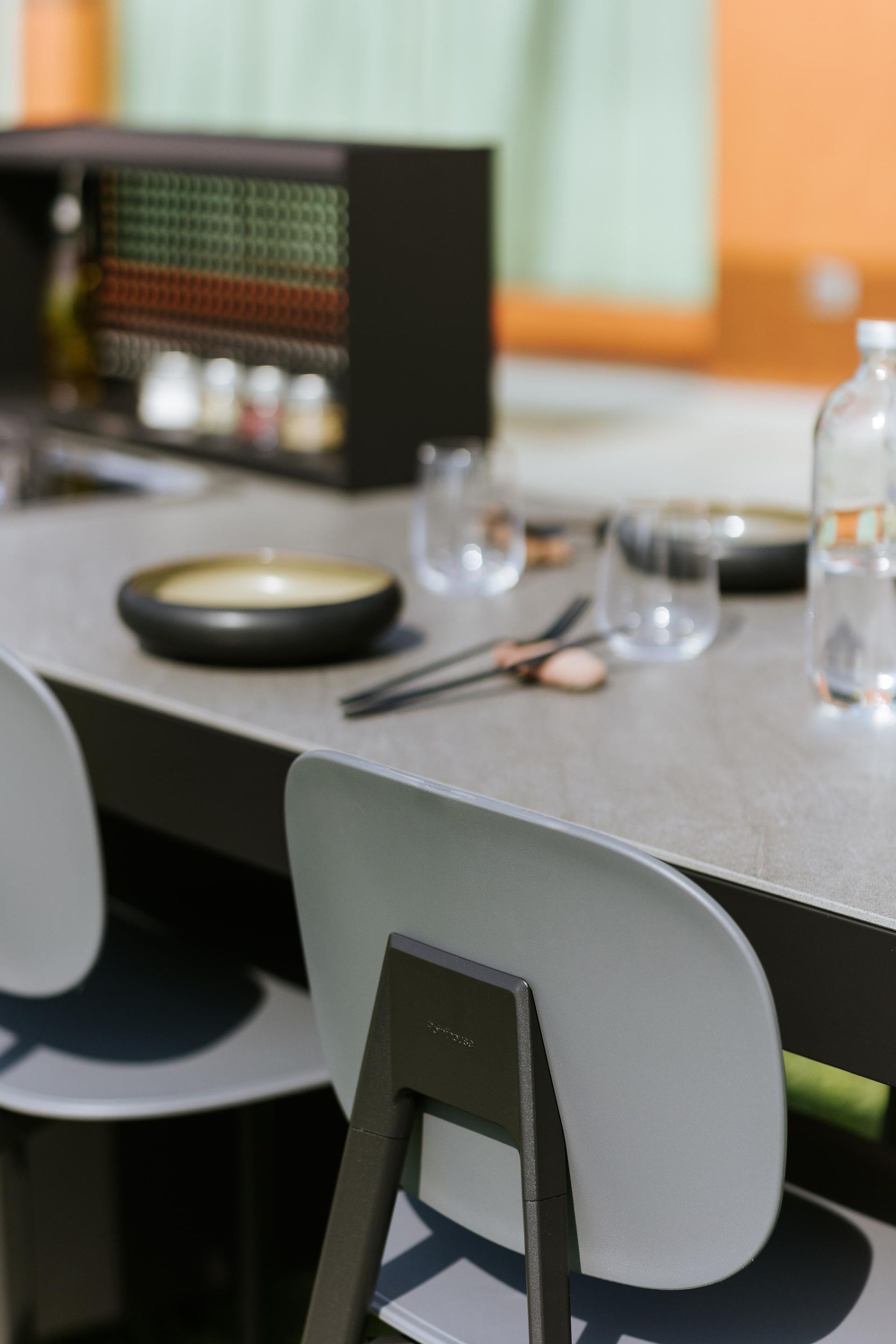 Sedie grigie attorno a un tavolo apparecchiato con stoviglie, vicino a un mobile scuro, con uno sfondo esterno.