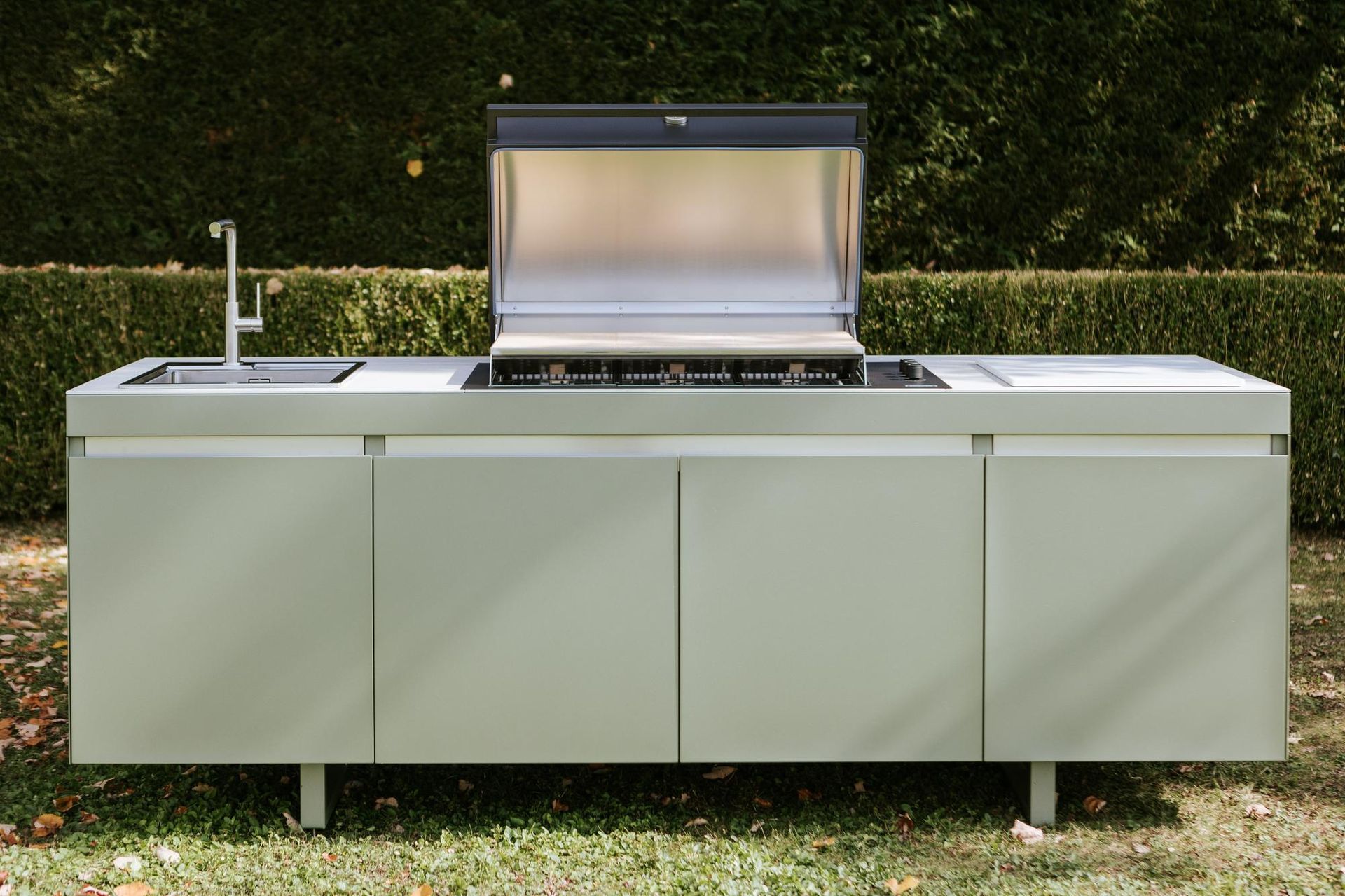 Cucina esterna con griglia, lavello e armadietti di fronte a una siepe.