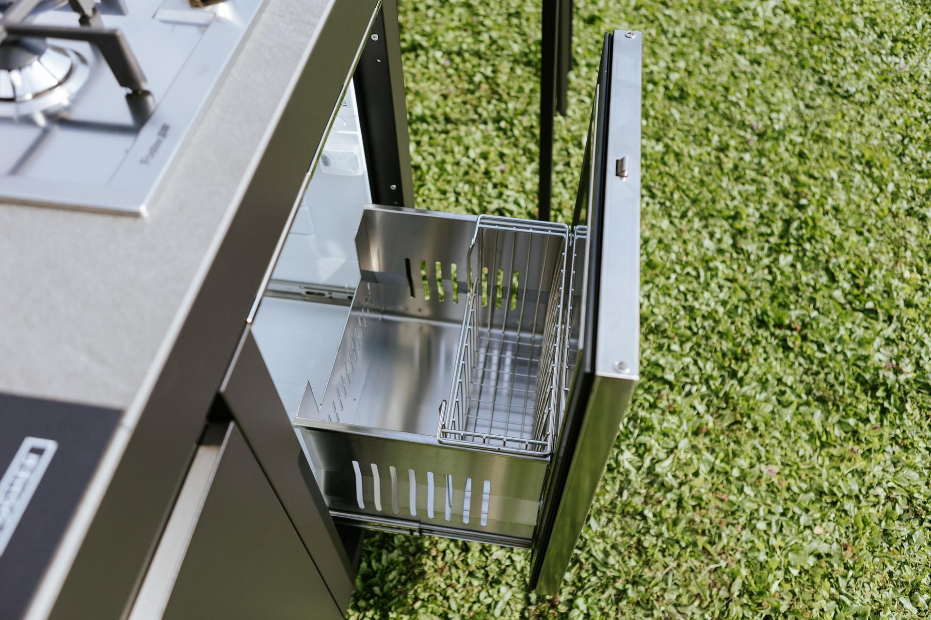 Cucina da esterno con cassetto aperto contenente un cestello portautensili in acciaio inox, posta su un prato verde.