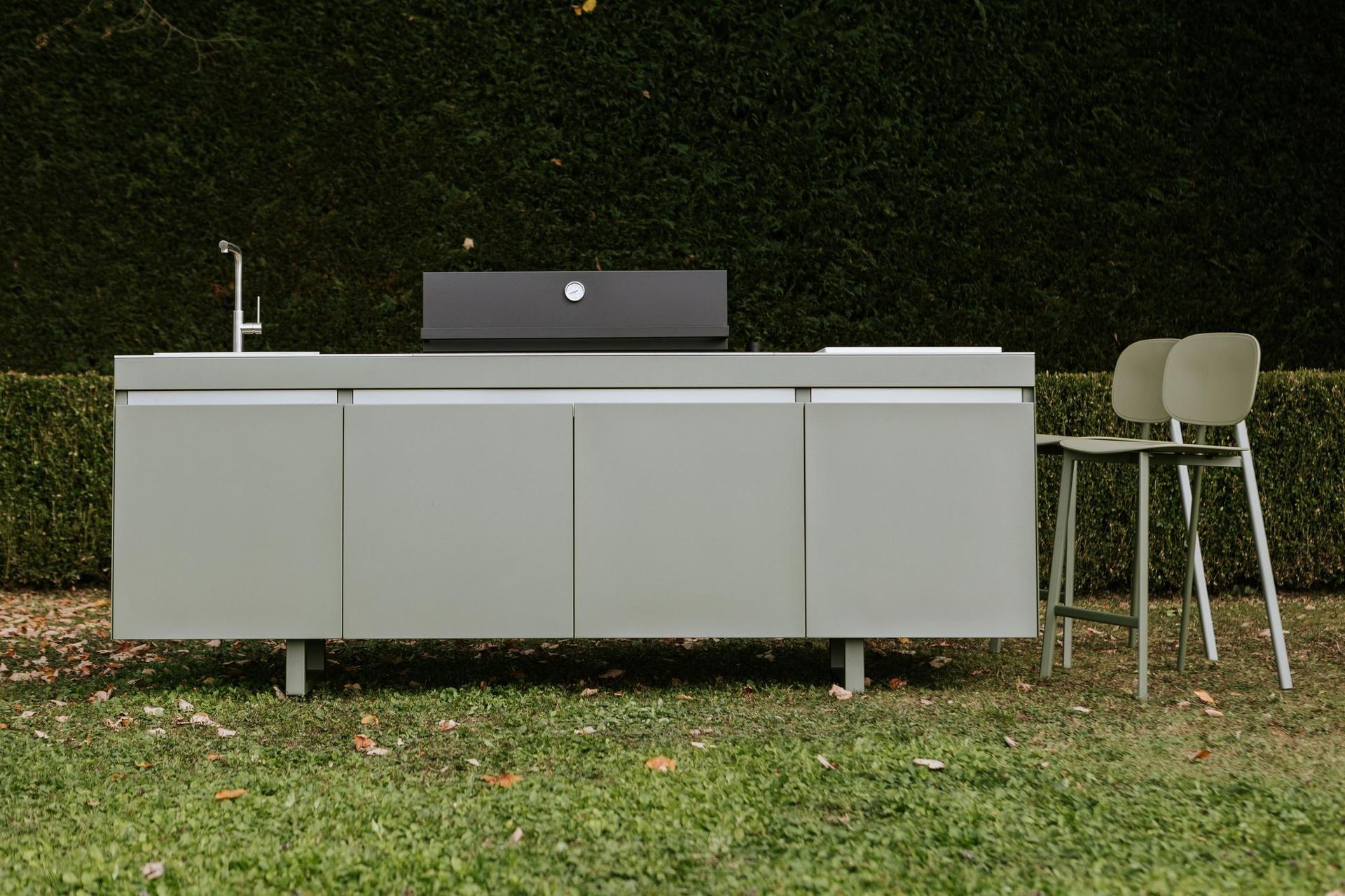 Cucina all'aperto con mobili verdi, lavello, griglia e uno sgabello su un prato.