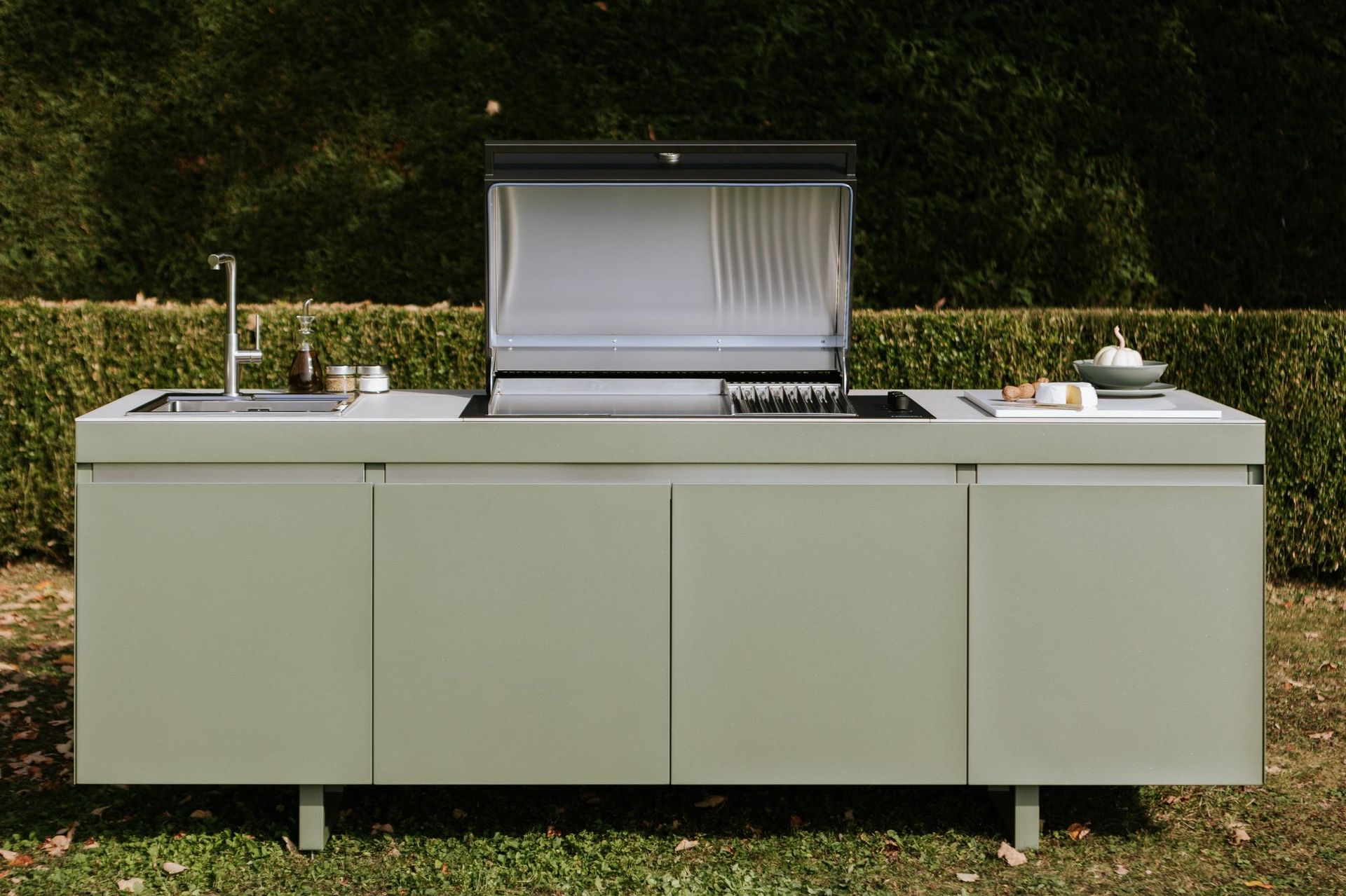 Cucina esterna con lavello, grill e spazio per preparare i pasti; mobili verde salvia, in un ambiente erboso.