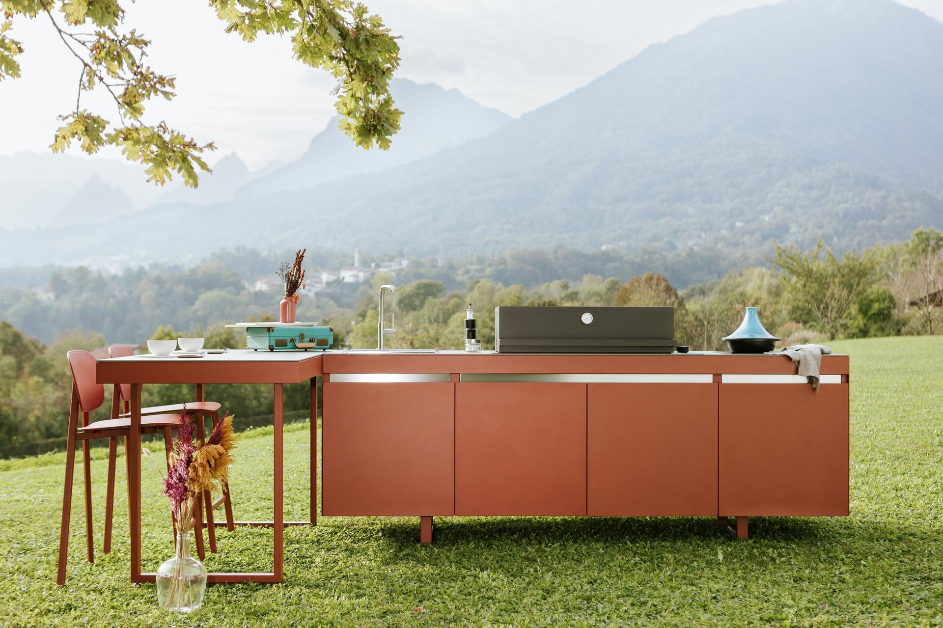 Cucina esterna con mobile color ruggine, tavolo e griglia sullo sfondo delle montagne.