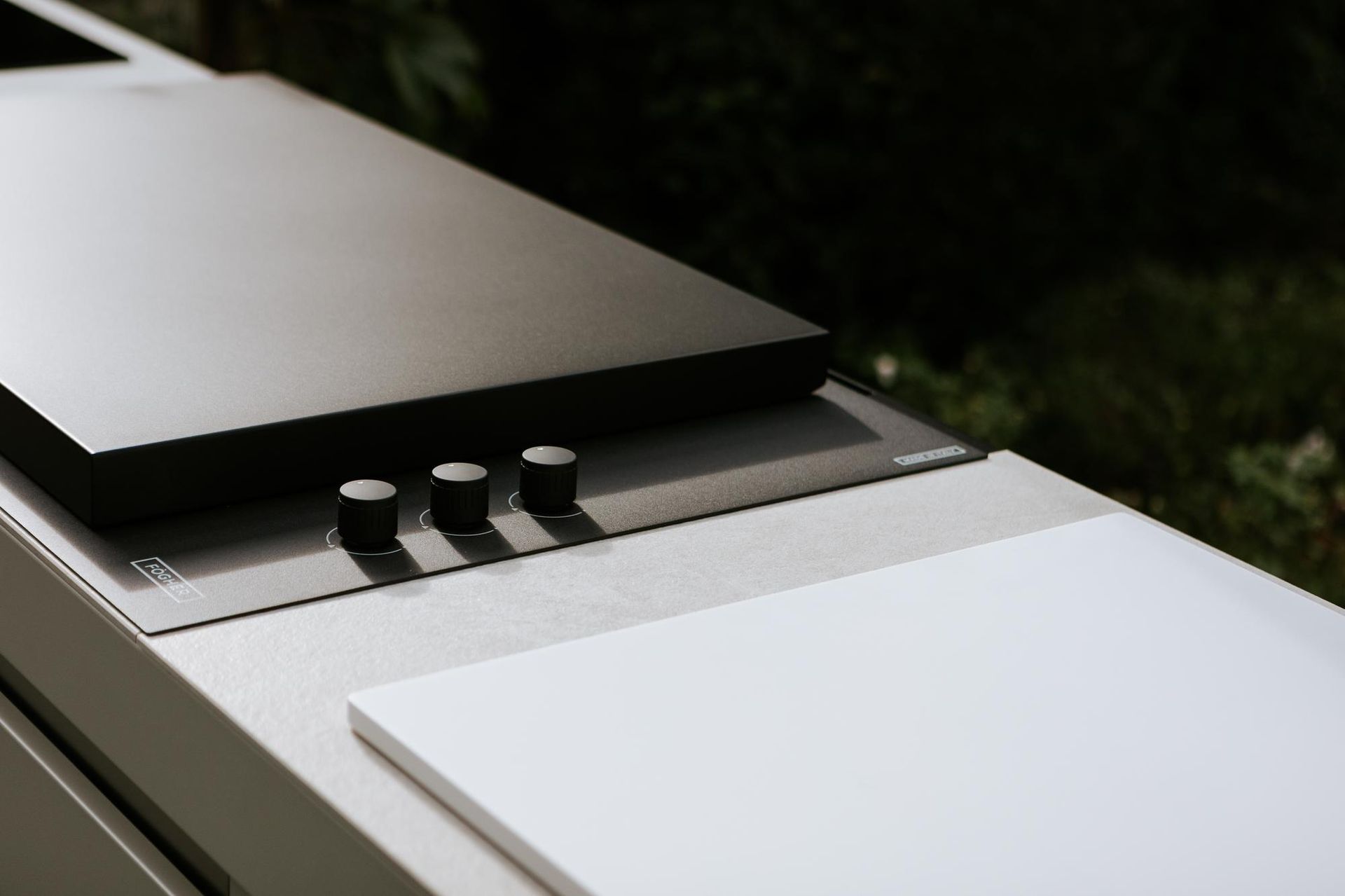 Stazione di cottura esterna con piano cottura nero, manopole e piano di lavoro bianco.