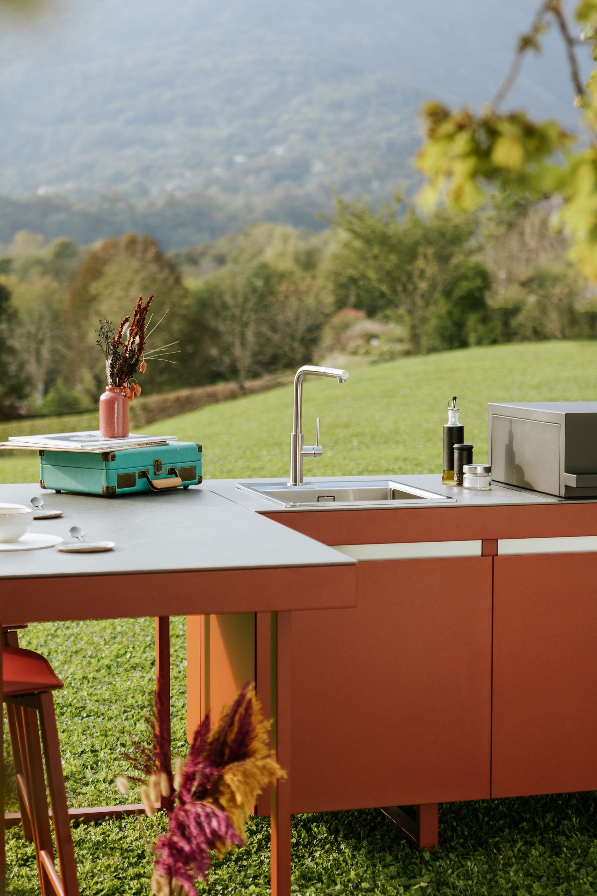 Cucina esterna con lavello, piano di lavoro e mobili, situata in un campo erboso con montagne sullo sfondo.