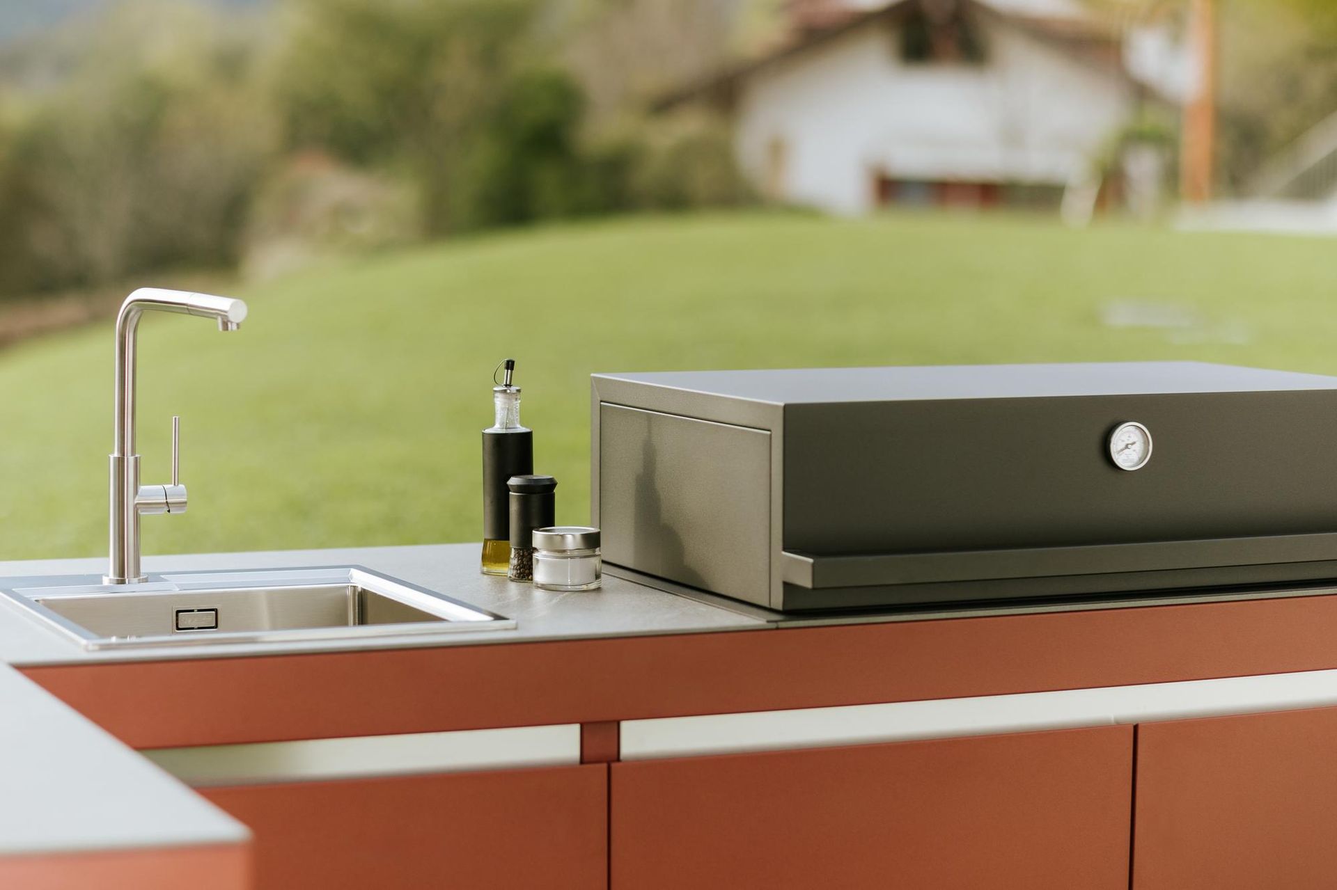 Cucina all'aperto con lavello, griglia e condimenti su un bancone, con sfondo naturale sfocato.