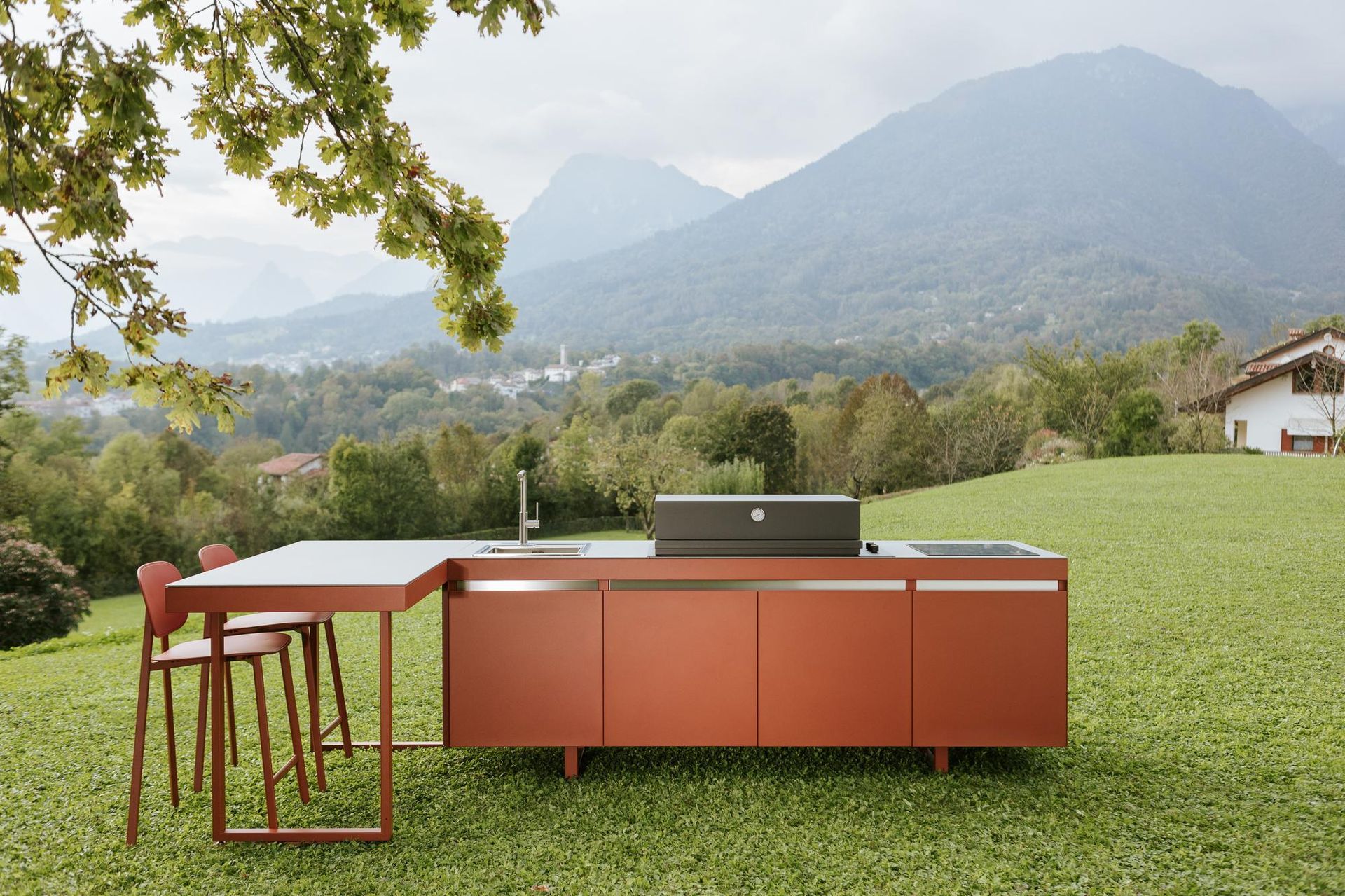 Cucina rossa all'aperto con lavello, grill e tavolo in un campo erboso con vista sulle montagne.