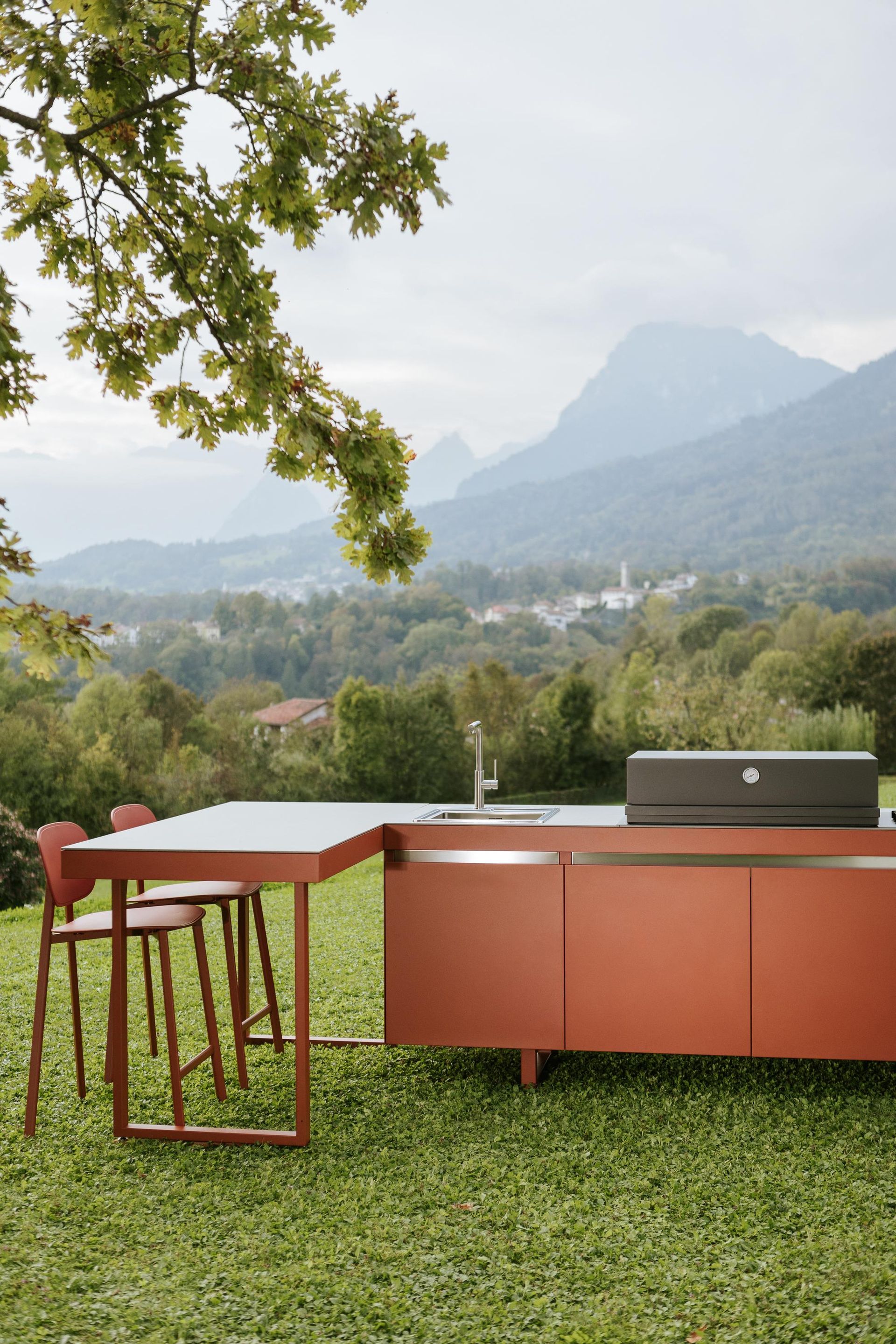 Cucina all'aperto con bancone e tavolo arancioni; montagne sullo sfondo, erba verde, due sgabelli da bar, cielo coperto.