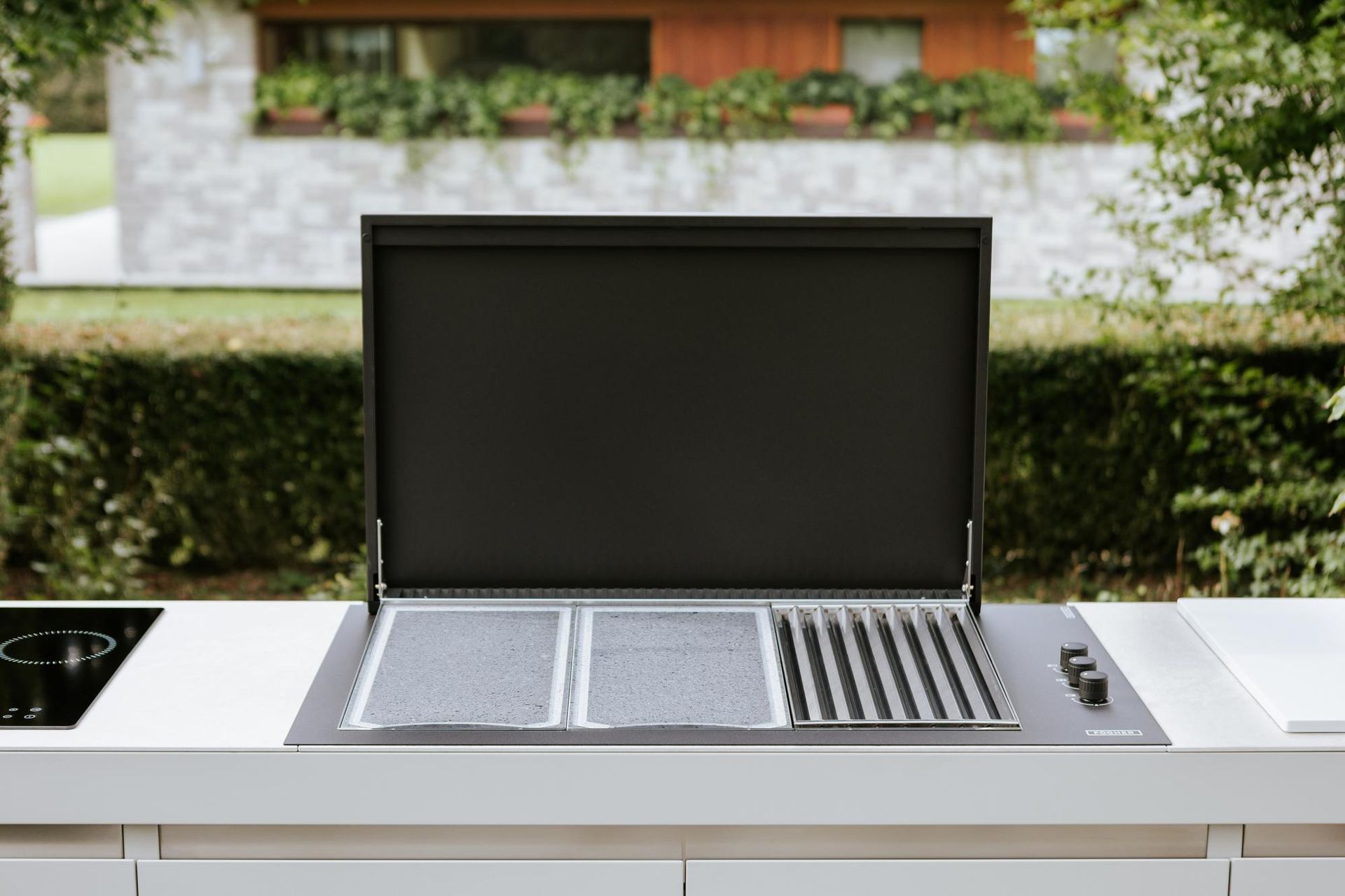 Griglia da esterno con coperchio aperto, inserita in una moderna cucina bianca da esterno.