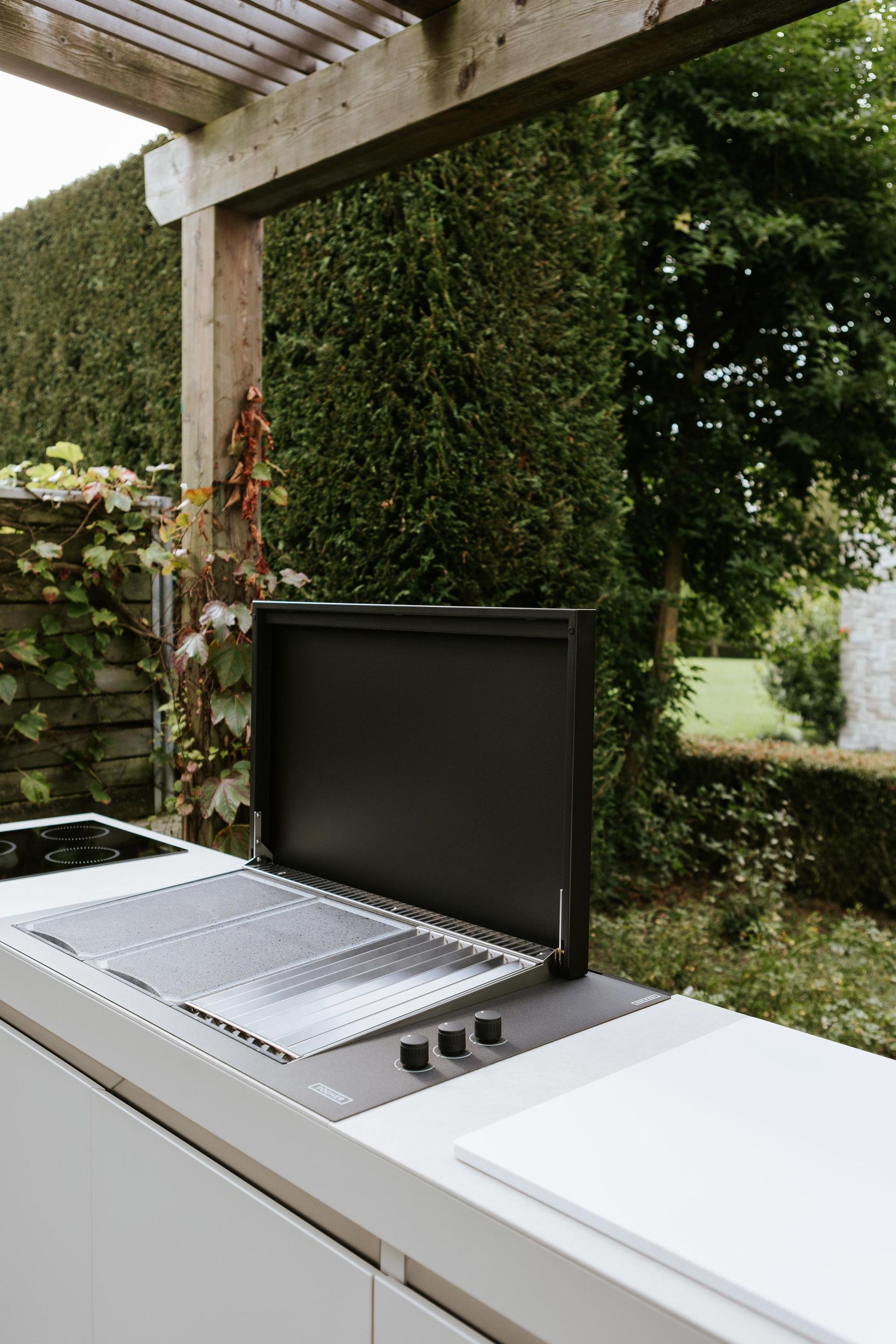 Cucina all'aperto con griglia sotto un pergolato in legno, sullo sfondo una rigogliosa siepe verde.