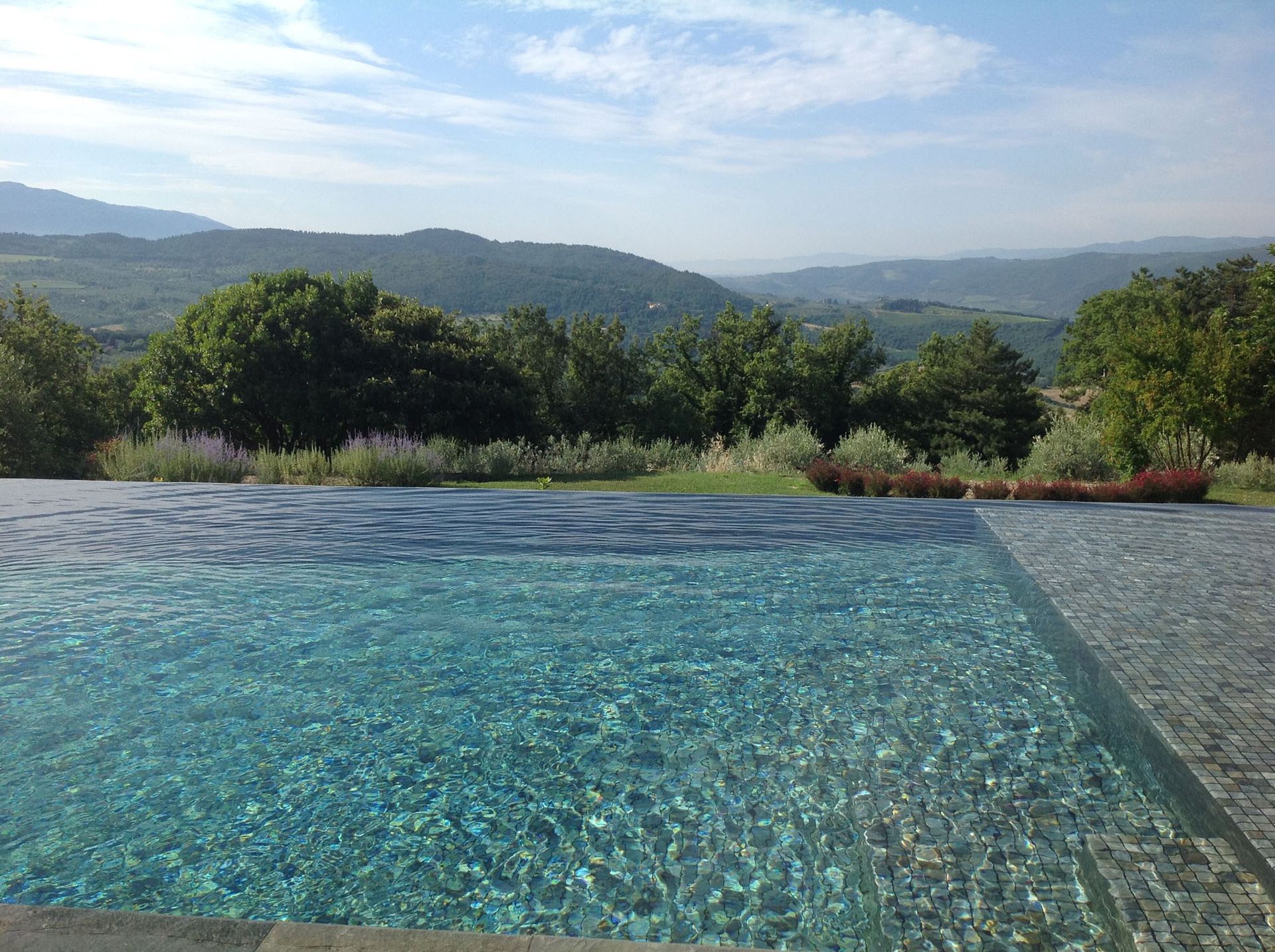 Piscina a sfioro con vista sulle verdi colline e montagne sotto un cielo azzurro.