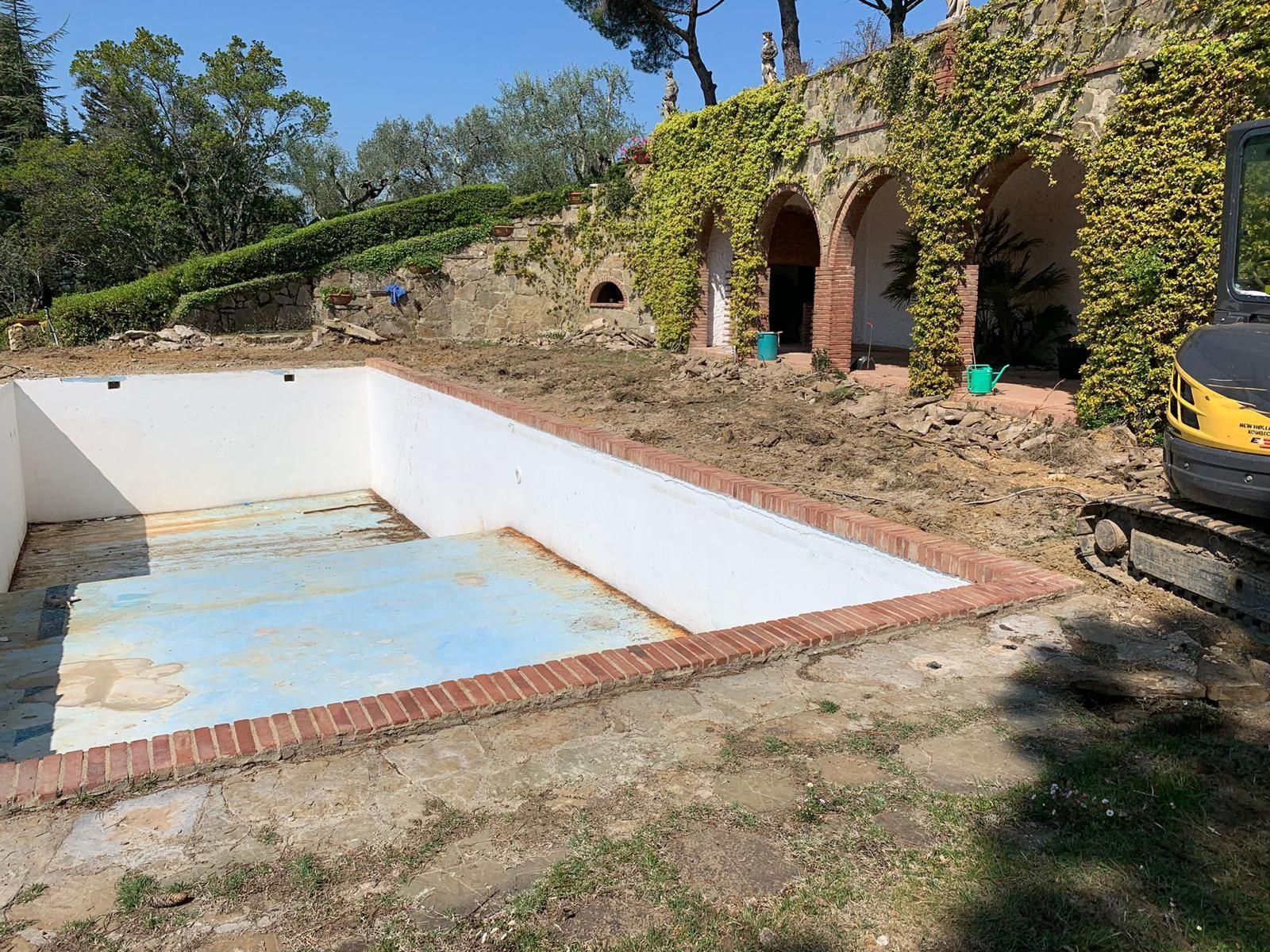 Piscina in costruzione con bordo in mattoni, terra battuta e un edificio con arcate ricoperte di edera.