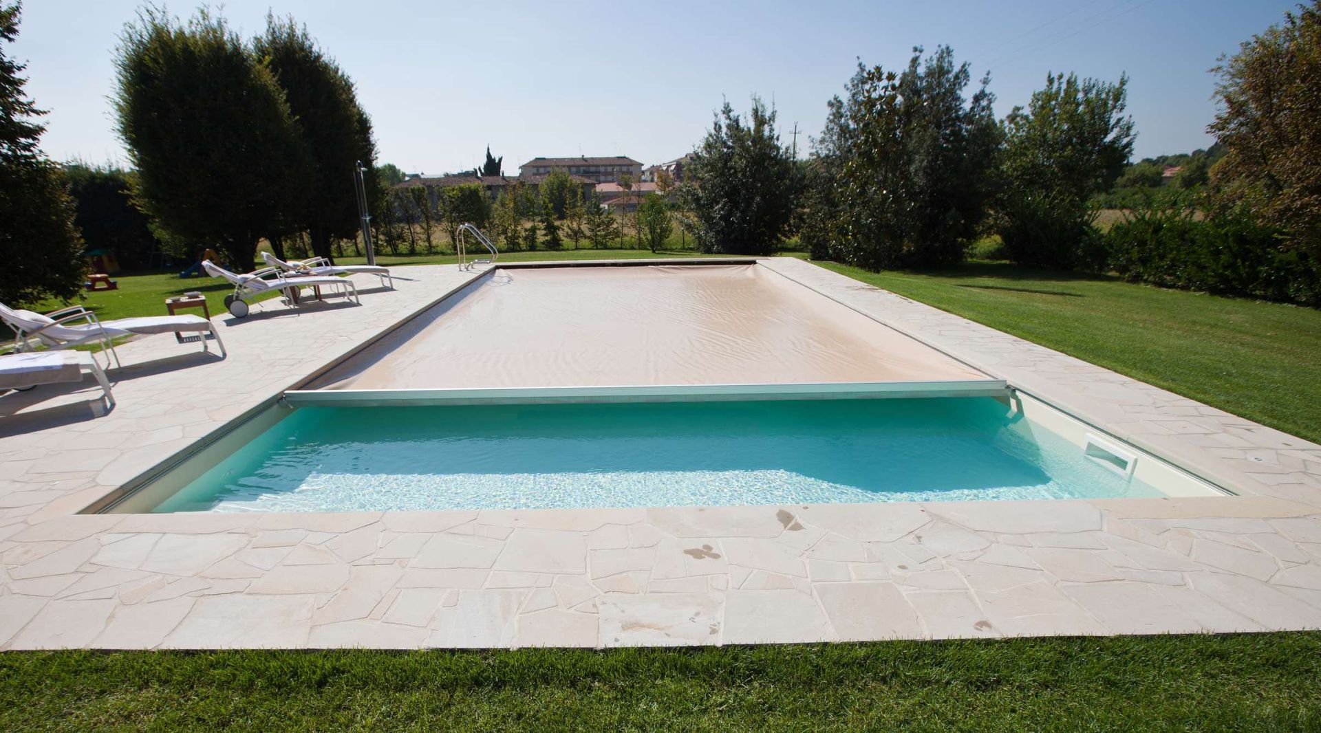 Piscina con superficie parzialmente coperta, circondata da pietra beige, erba e alberi.