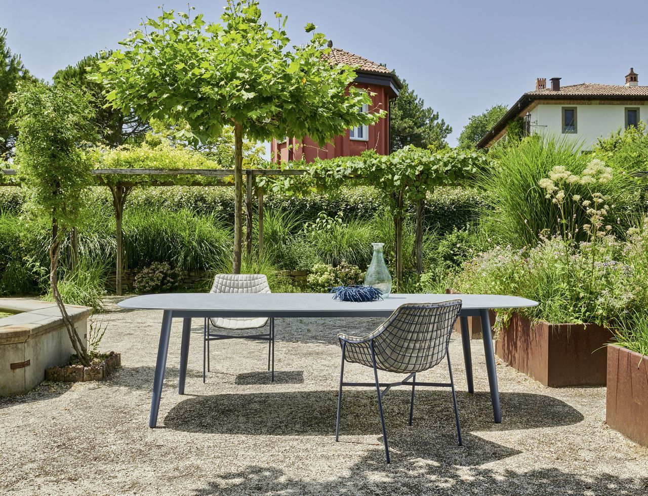 Tavolo da pranzo e sedie all'aperto su un patio in ghiaia circondato da vegetazione lussureggiante, alberi ed edifici.