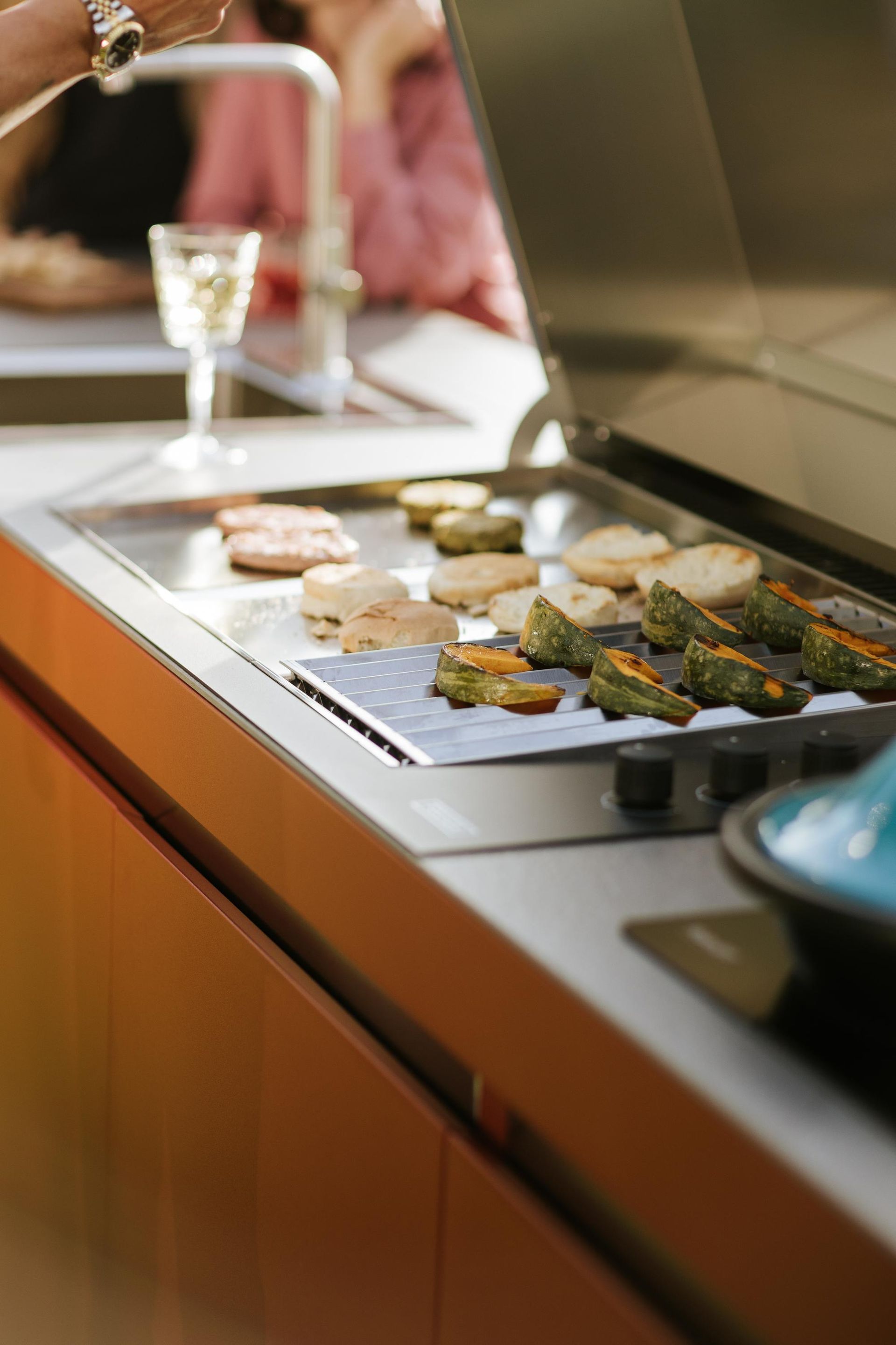 Griglia con vari alimenti in cottura, appoggiata su un mobiletto arancione. Un bicchiere di vino è lì vicino.