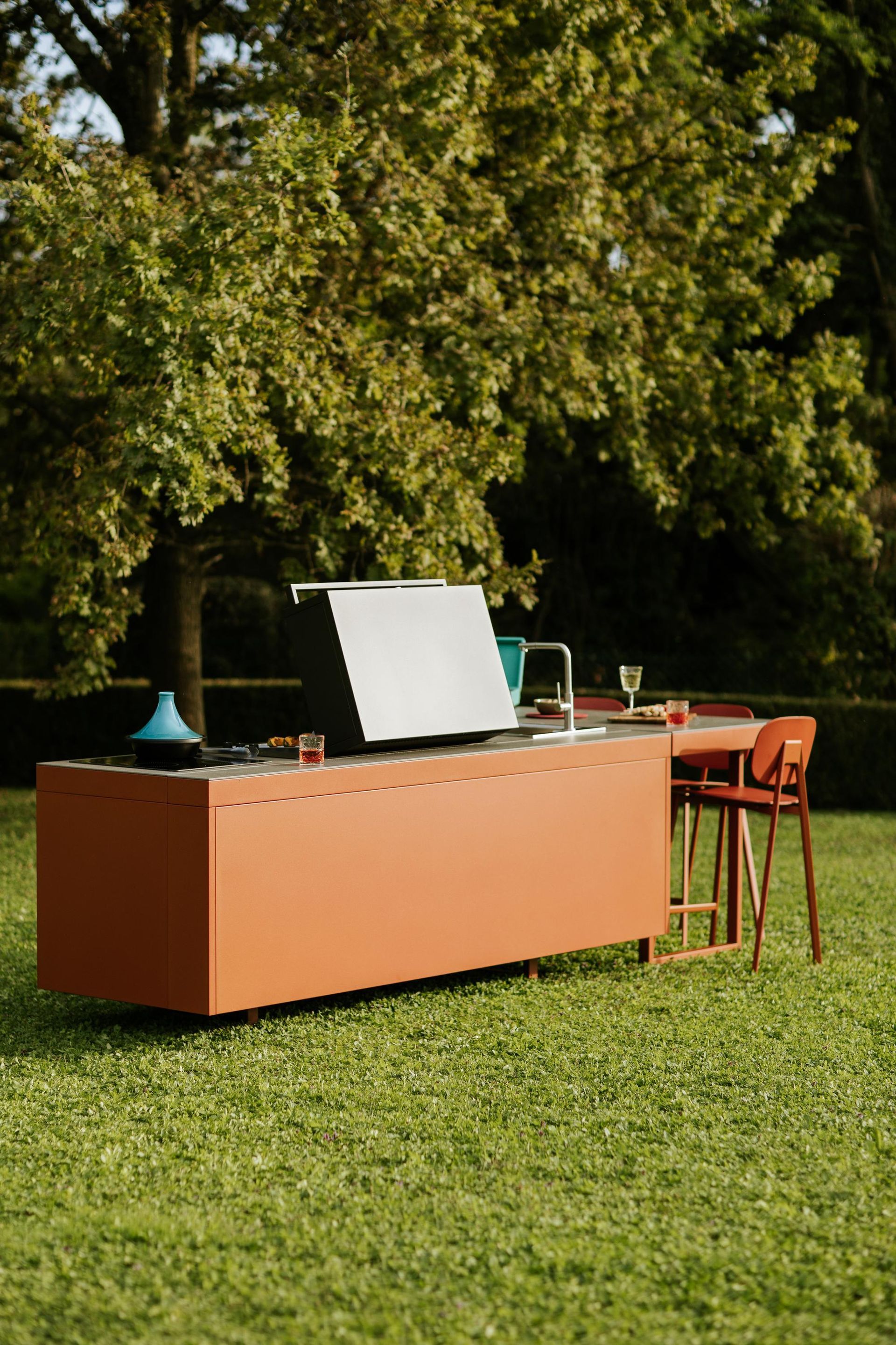 Cucina arancione da esterno con griglia, lavello e sgabello da bar in un cortile erboso, con alberi sullo sfondo.