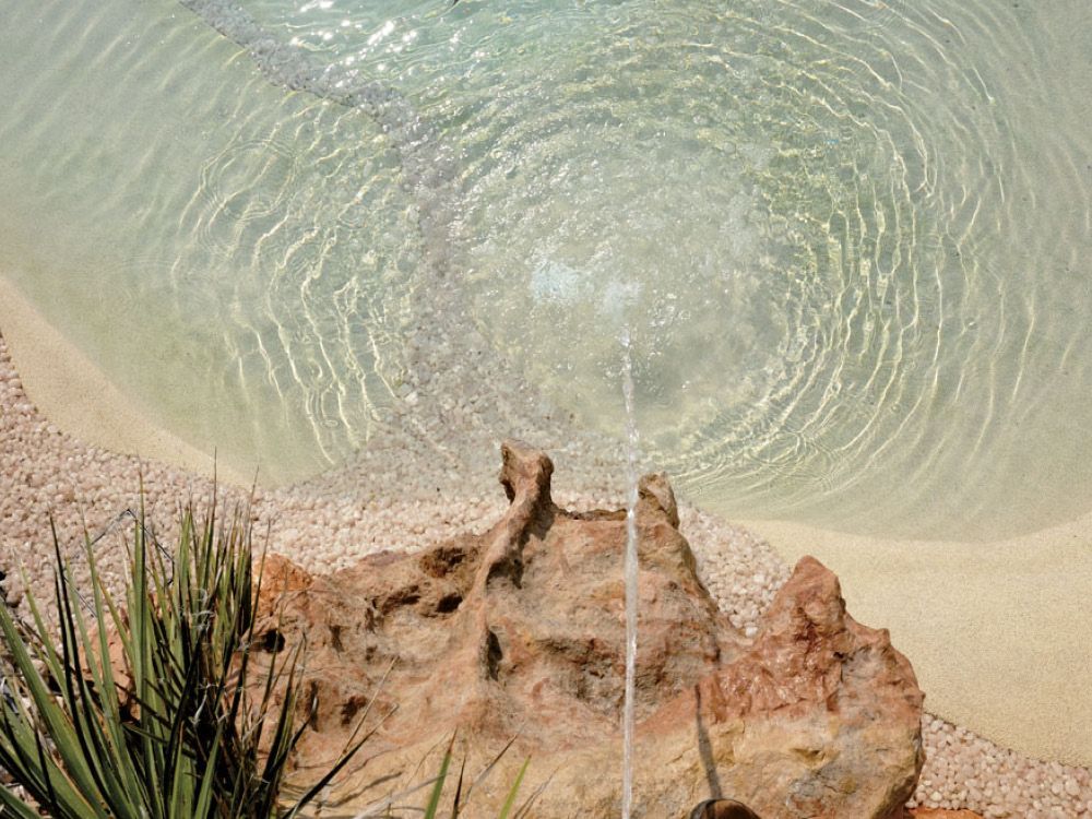 Fontana con getto d'acqua che crea increspature in una piscina turchese, con bordo roccioso e vegetazione.