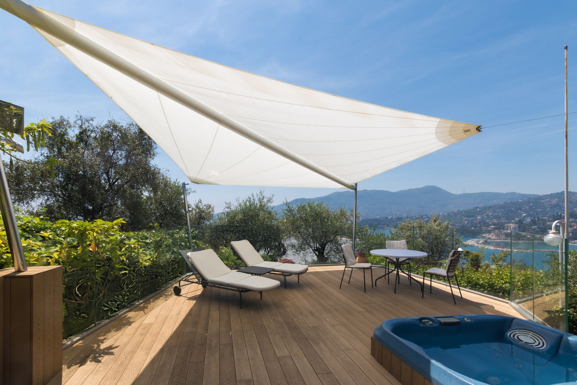 Terrazza in legno con sedie a sdraio, tavolino e vasca idromassaggio sotto un ombrellone con vista sull'acqua.