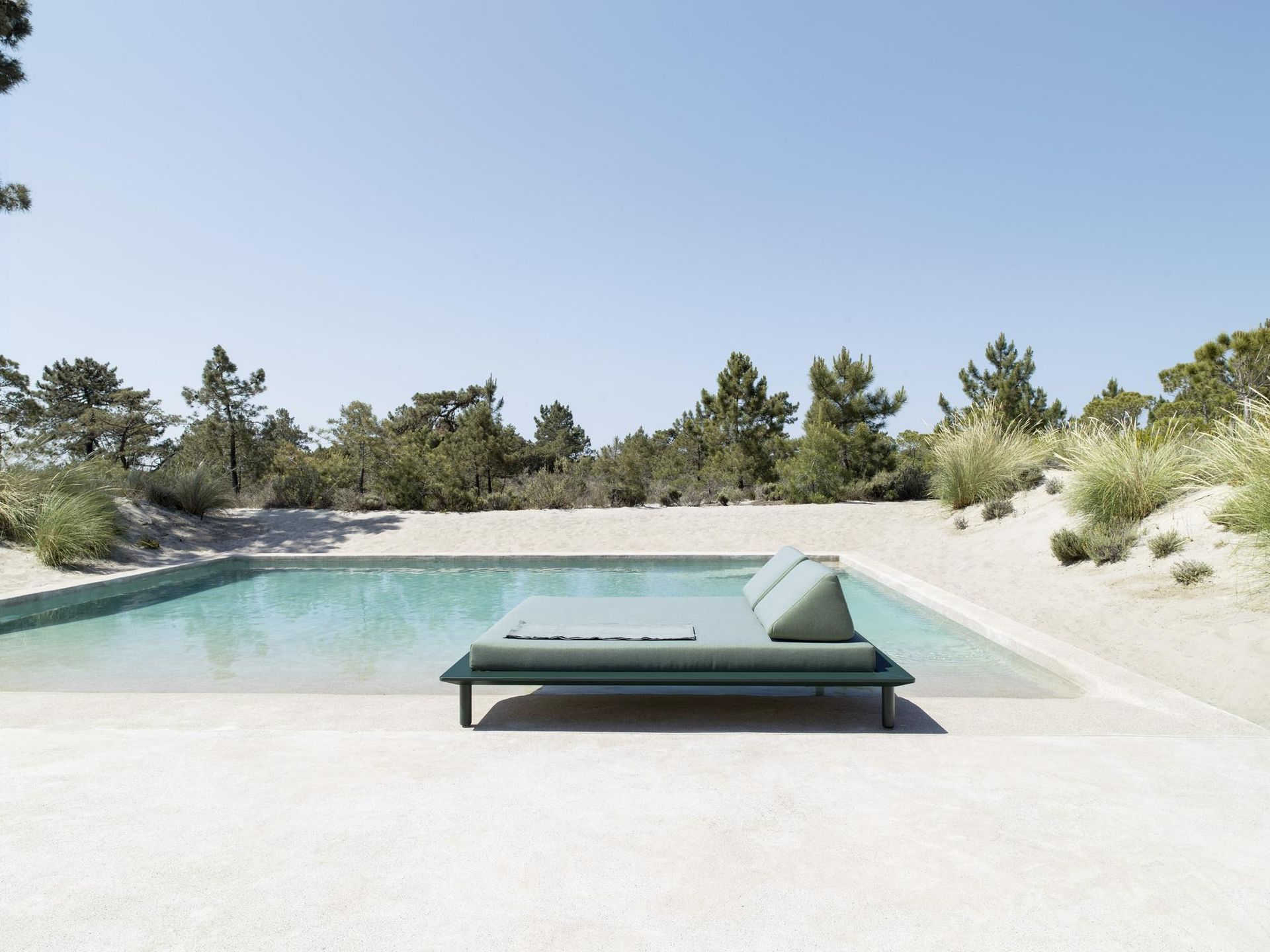 Chaise longue a bordo piscina, accanto a una piscina turchese circondata dal verde e sotto un cielo limpido.